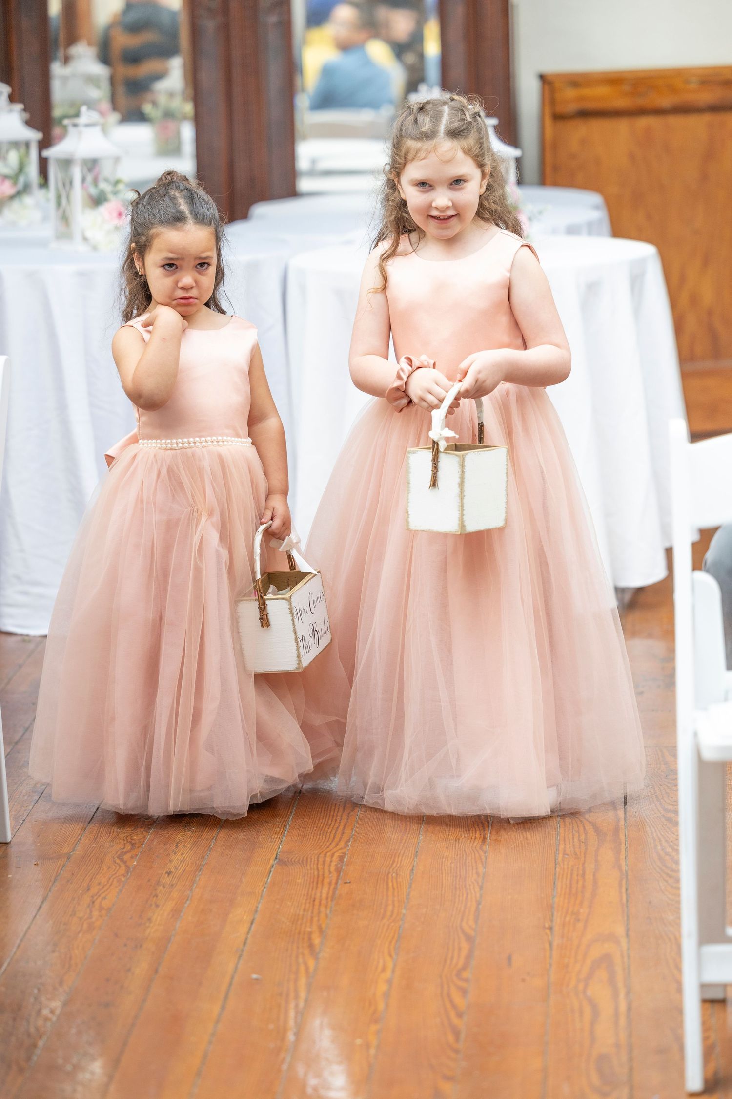 Young flower girls in blush pink tulle dresses walk down a wooden aisle at an elegant wedding ceremony.