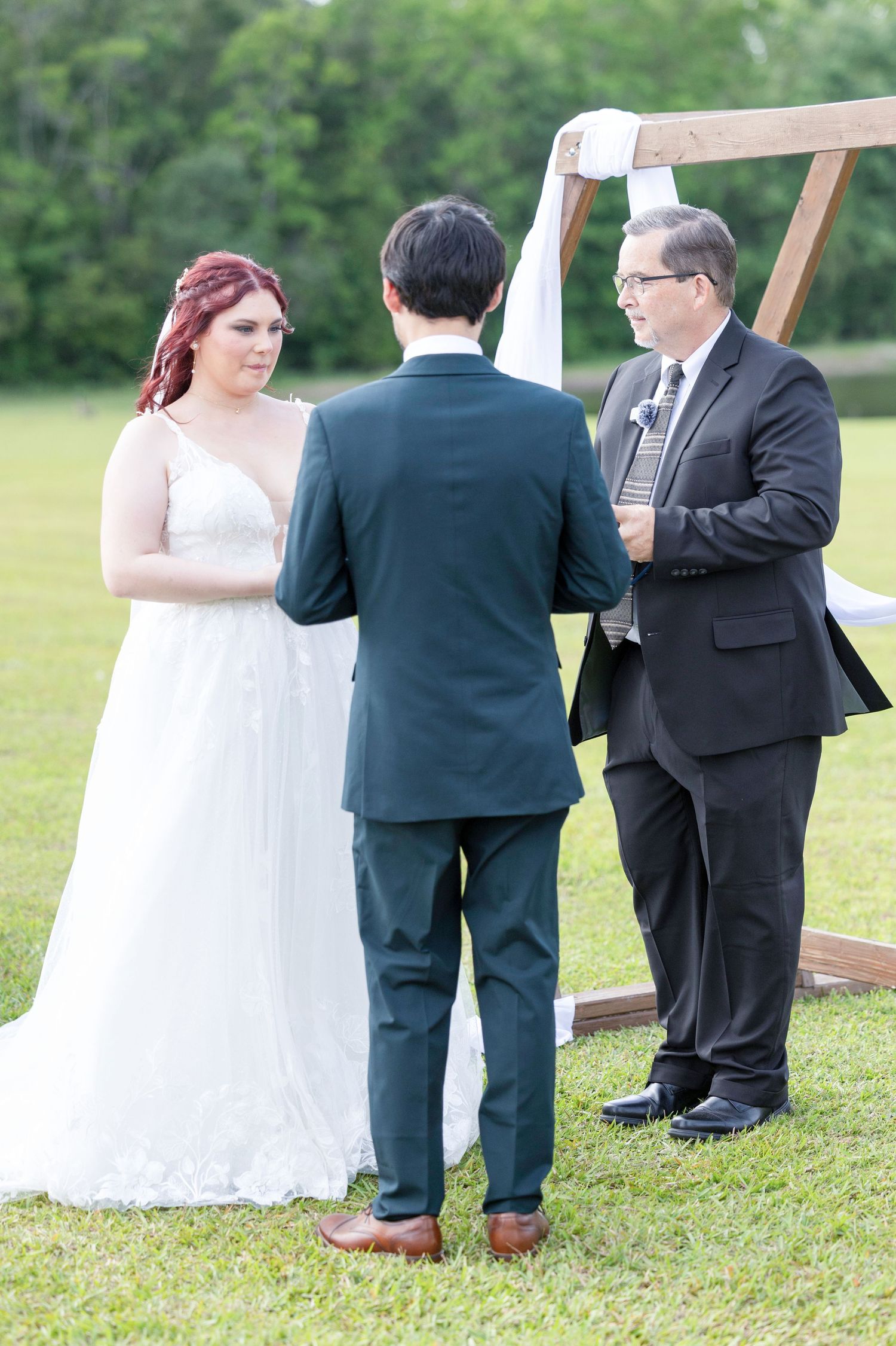 Outdoor wedding ceremony shows officiant and couple standing together on grassy lawn during daytime service.