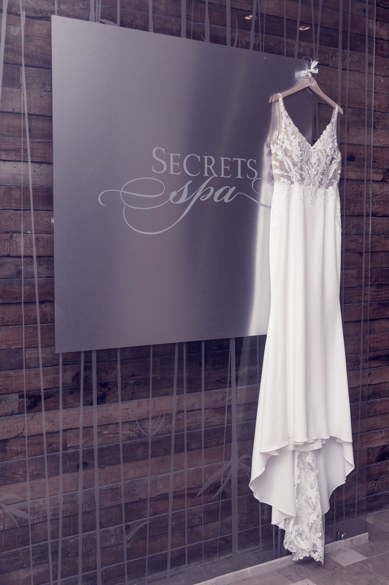A white wedding dress hangs elegantly against a wall with a 'Secrets Spa' sign in muted lighting.