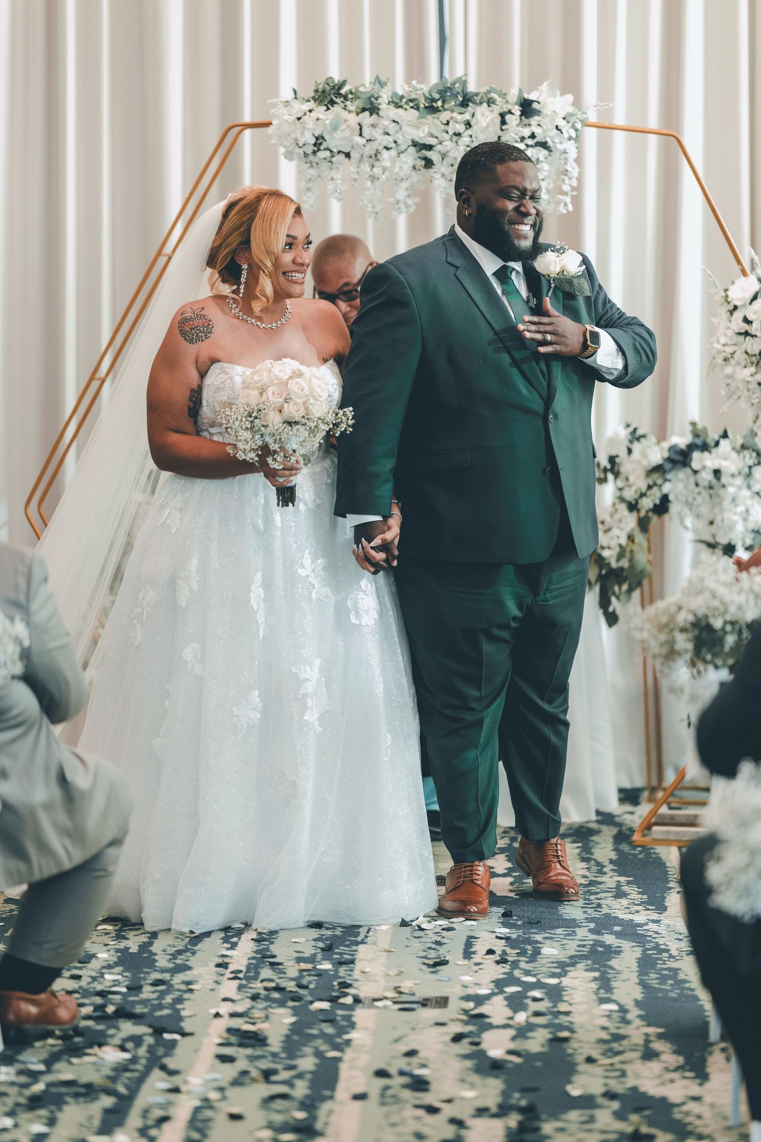 A joyful couple shares a moment during their wedding ceremony under a copper arch decorated with white flowers.