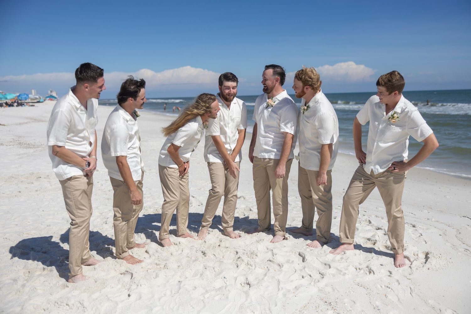 Wedding party wearing khaki pants poses playfully on a sunny white sand beach with ocean waves in background.