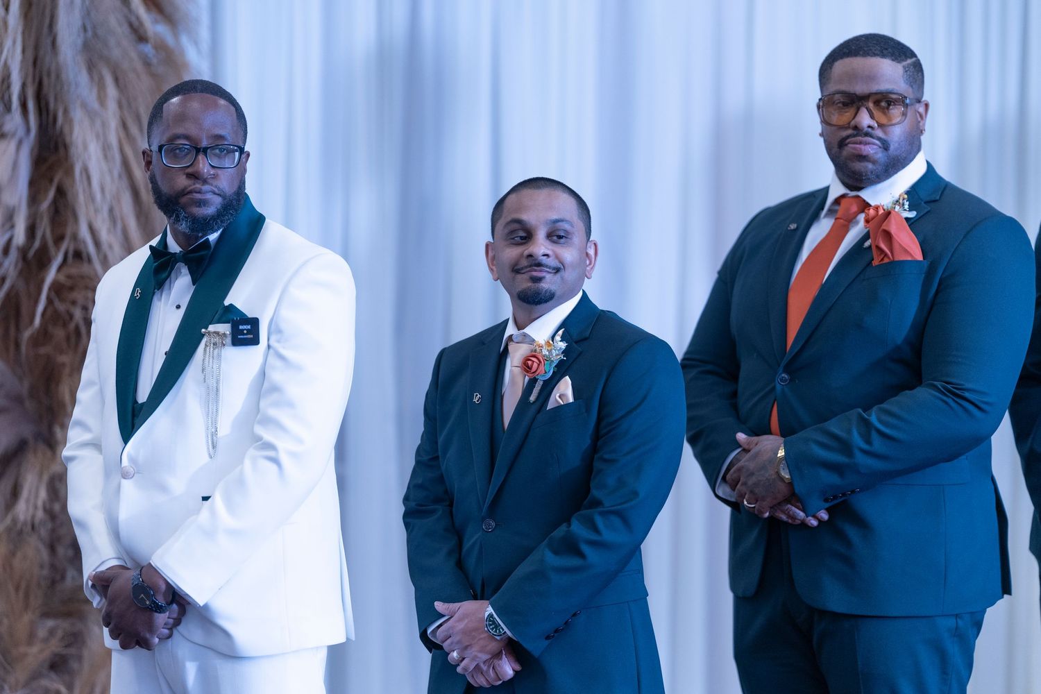 Three men in formal attire stand together at a wedding, wearing white, navy and blue suits with coordinating bow ties.