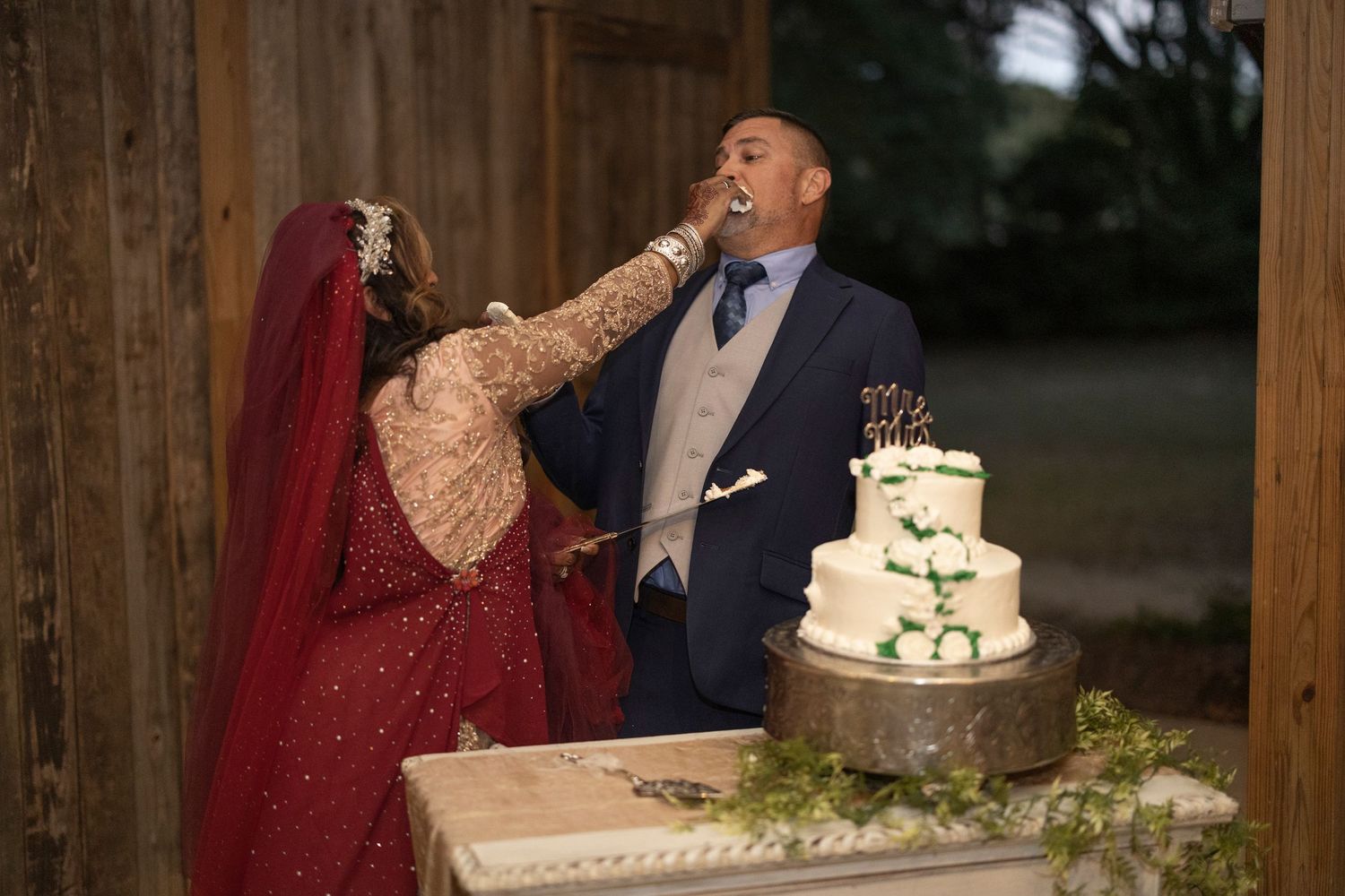 A couple stands next to a three-tiered wedding cake with white frosting and greenery decorations at a rustic venue.