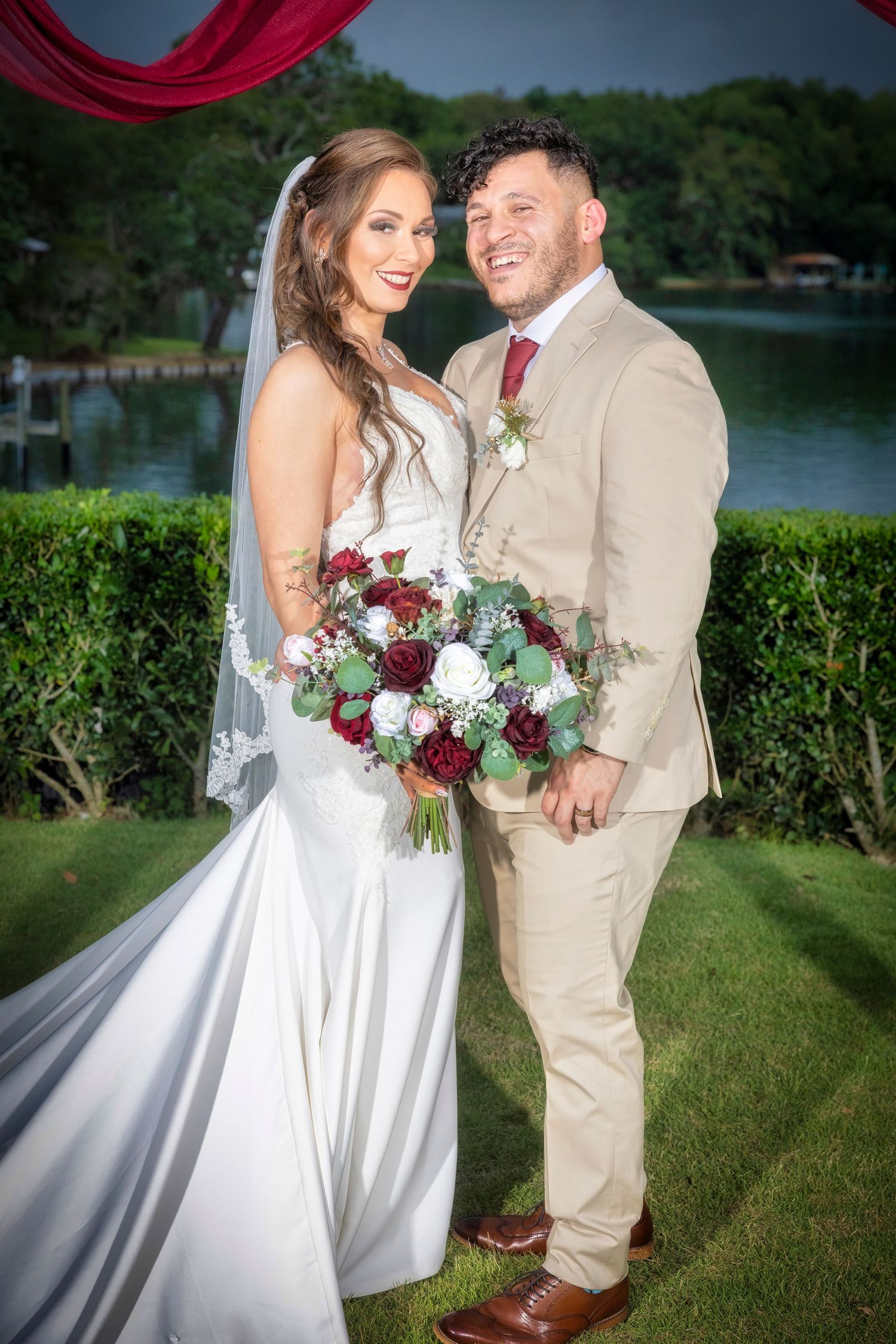 Newlyweds pose together outdoors with burgundy and white bouquet by a lakeside garden.