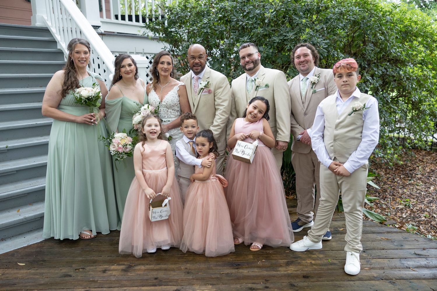 Wedding party poses on wooden deck wearing sage green and blush pink dresses with tan suits for outdoor celebration.