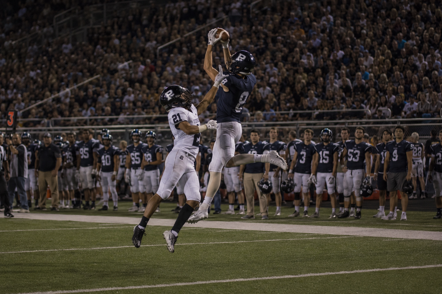 Football player #2 leaps to catch a pass over a defender during a night game on a well-lit field.