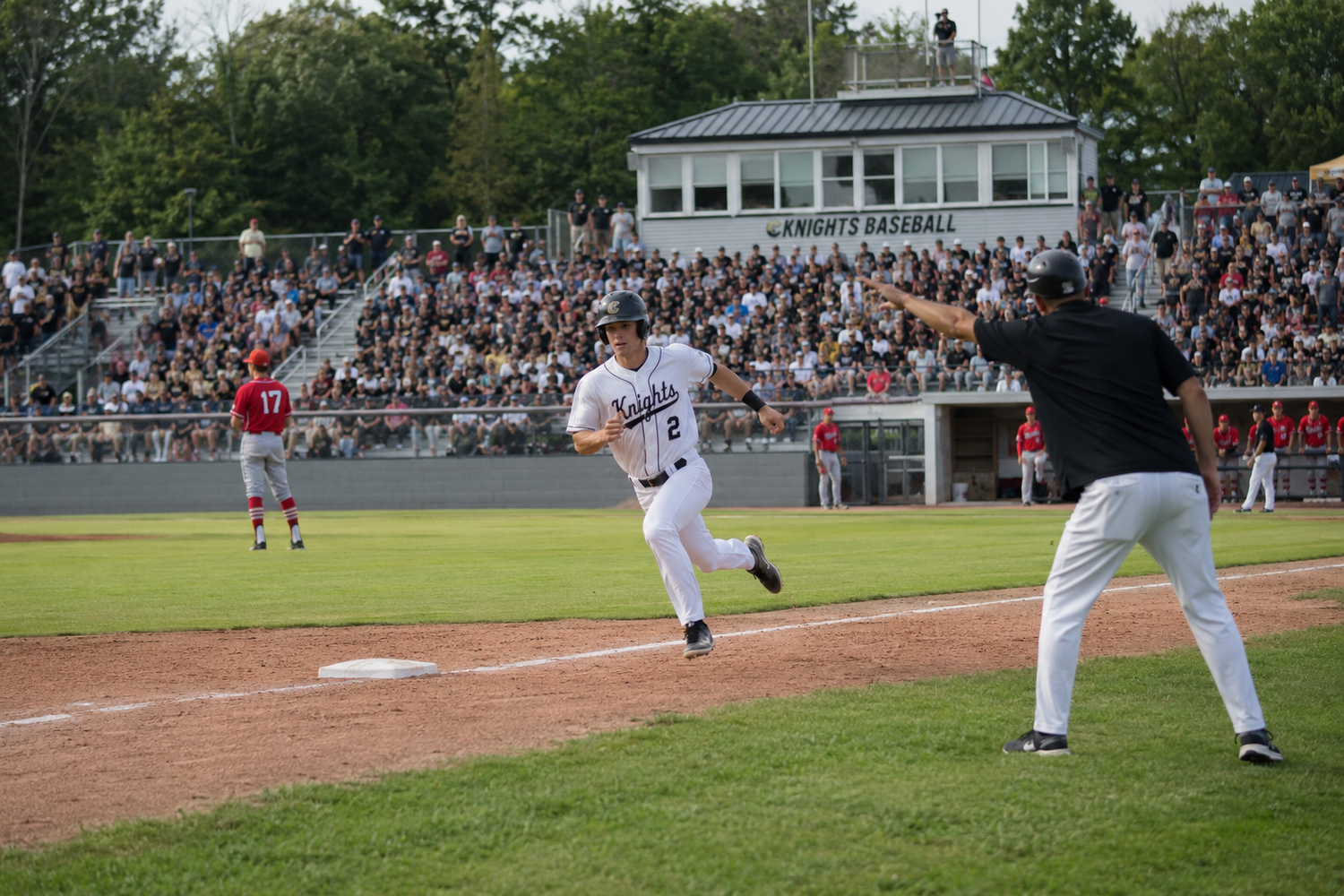 Baseball player rounds third base as coach signals during a packed high school stadium game.