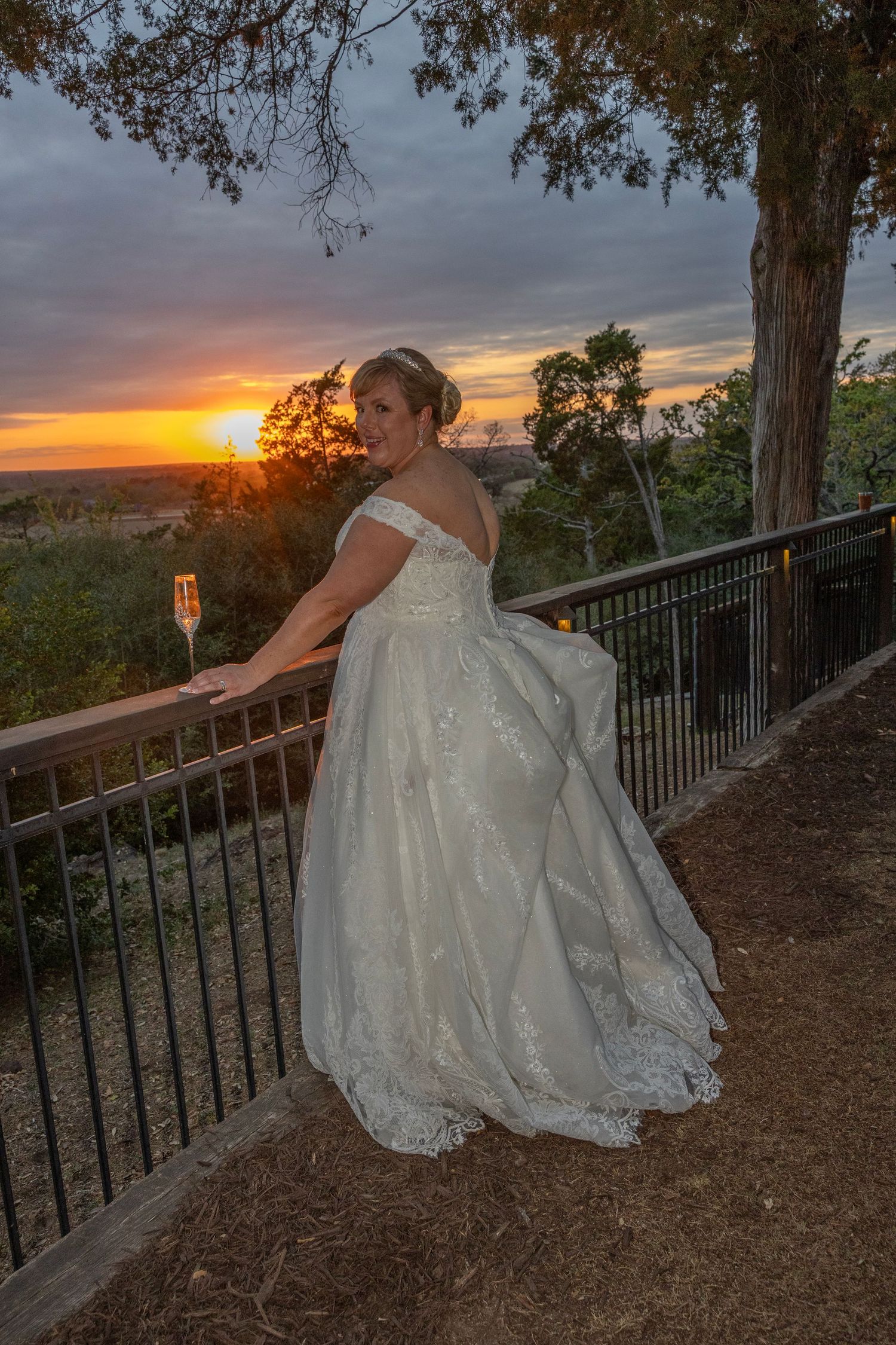 Bride in white gown holding champagne at sunset overlook, golden sky backdrop.