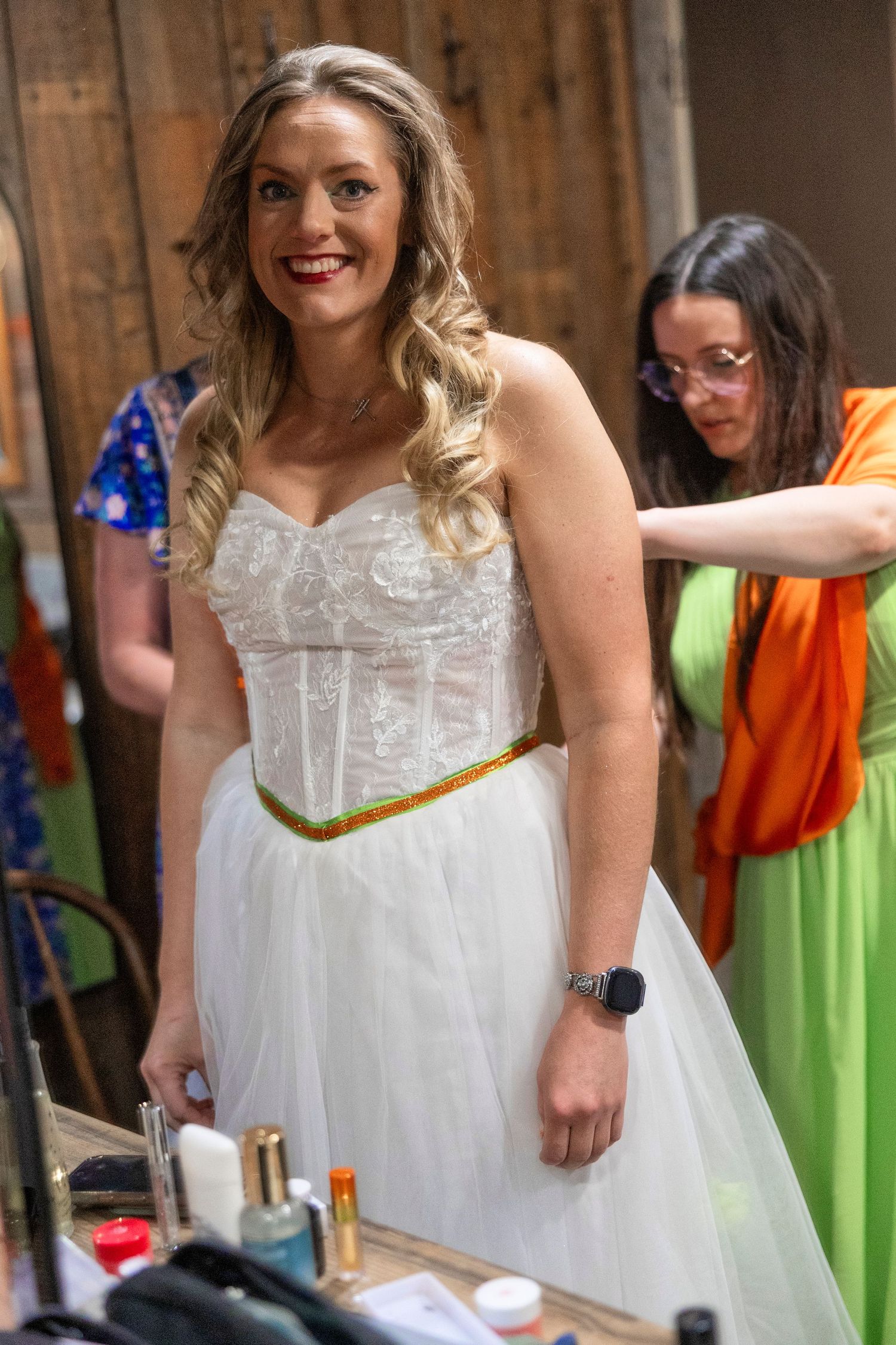 Bride in white lace corset wedding dress with gold belt smiles as bridesmaid in green adjusts her gown.