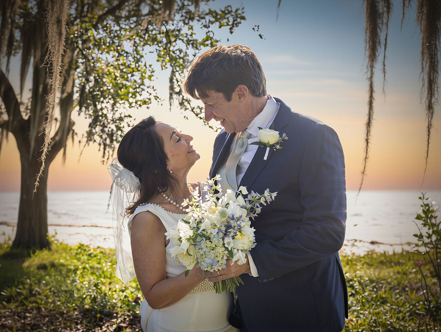 Bride and groom share a romantic moment at sunset near the ocean, with Spanish moss and a white floral bouquet.