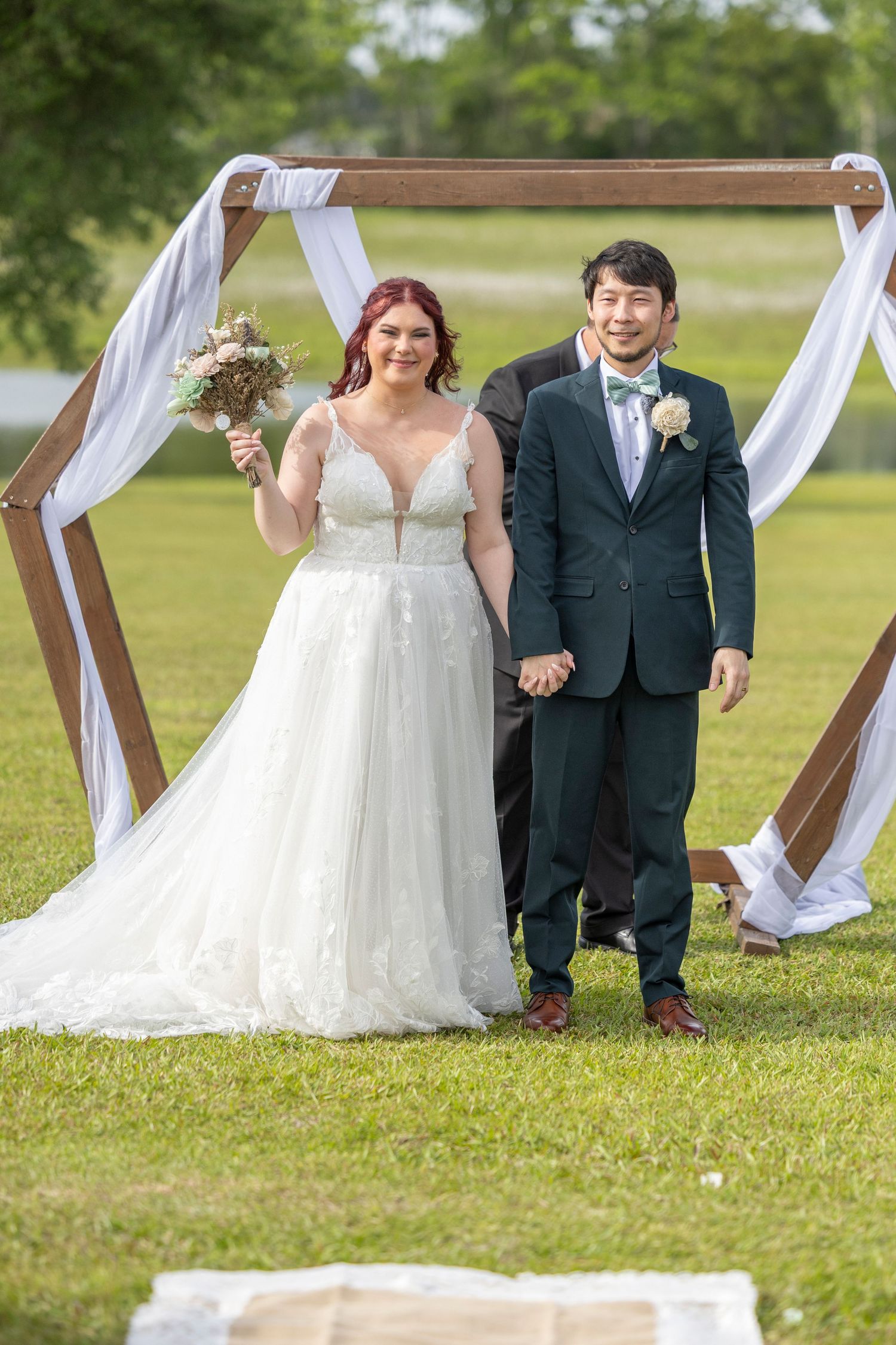 Wedding couple stands by rustic wooden arch draped with white fabric on a grassy lawn during outdoor ceremony.