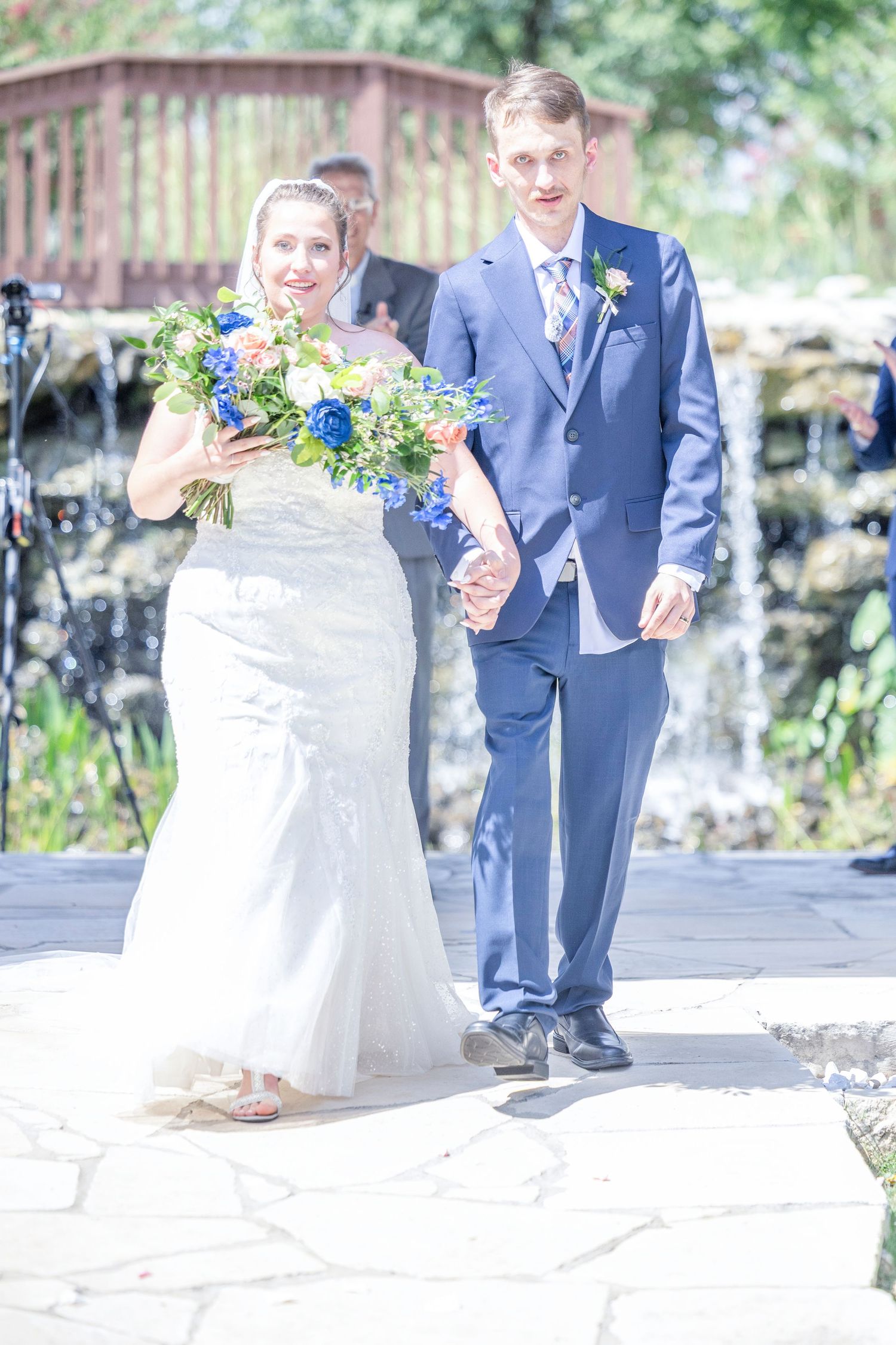 Newlywed couple in navy suit and white dress pose together outdoors holding colorful bouquets.