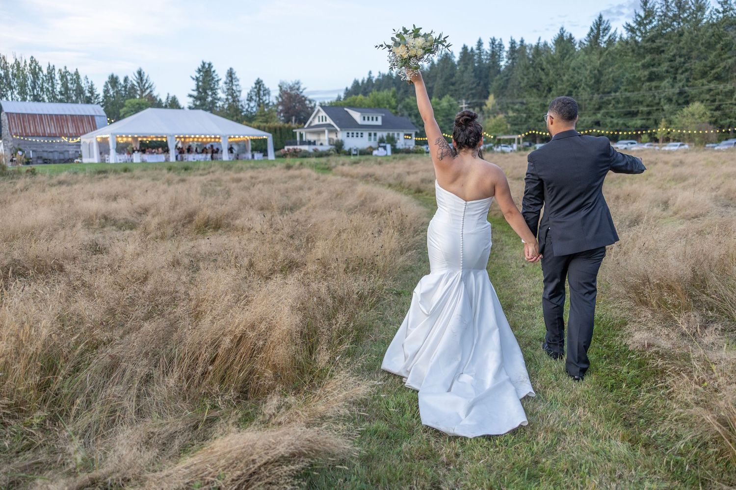 A couple in wedding attire walks through a scenic field with evergreen trees in the background.
