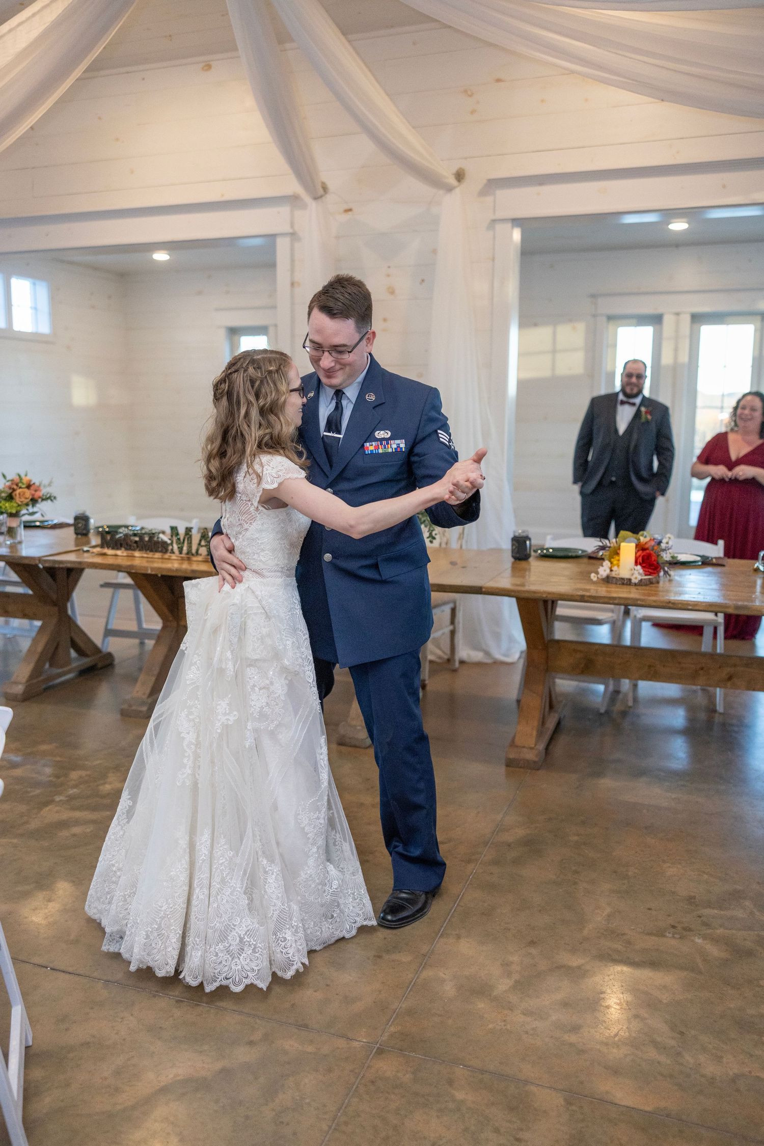 A couple shares their first dance at a wedding reception in a rustic venue with white walls and wooden beams.