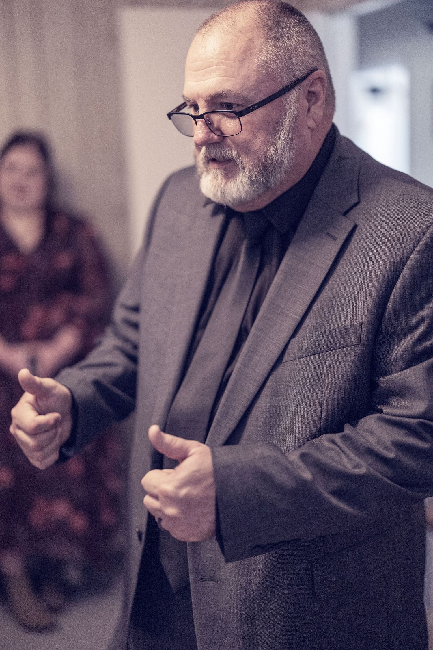 Professional wearing a gray suit and glasses gestures while speaking in an office environment.