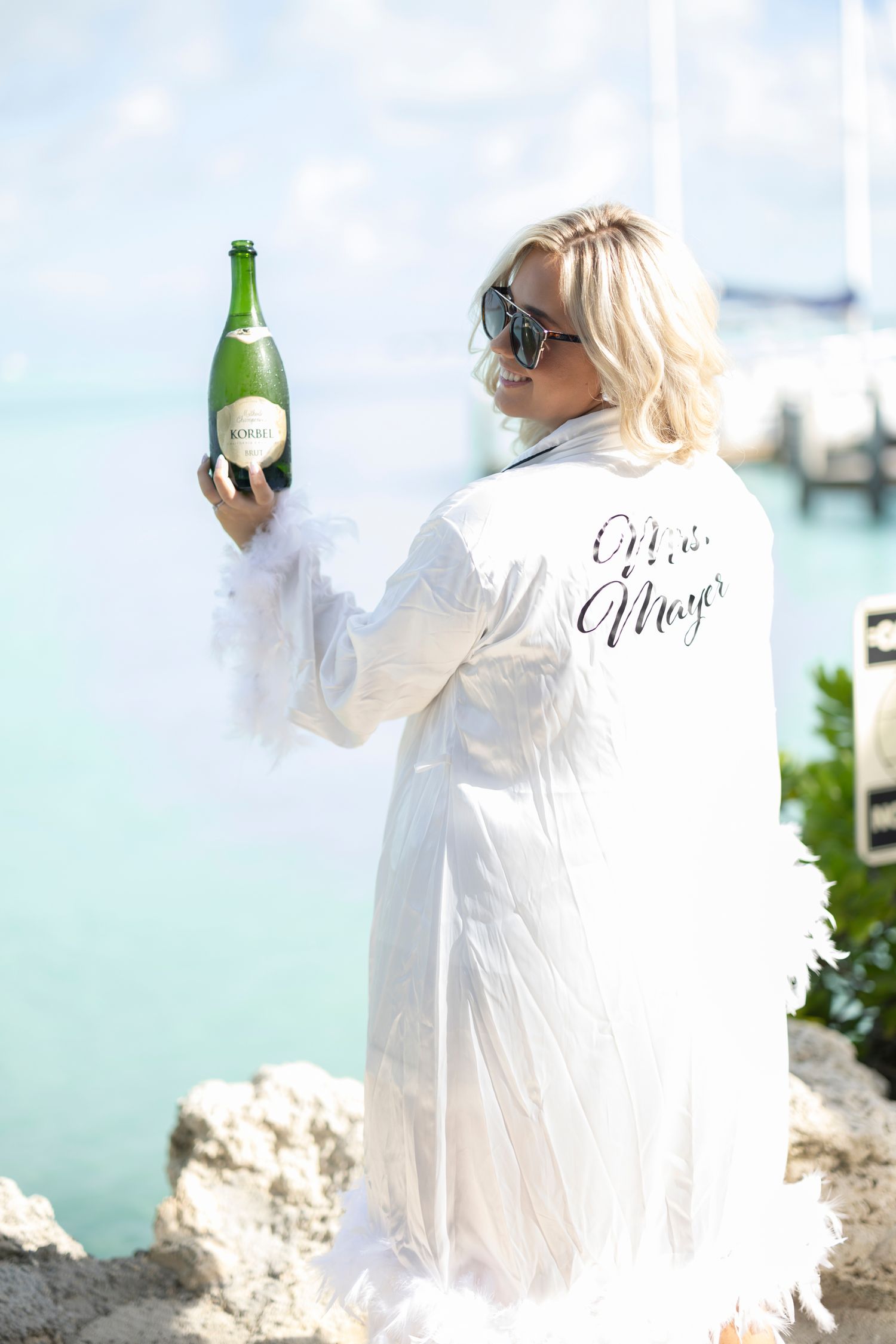 Woman in flowing white dress holds champagne bottle on rocky beach with turquoise water in background.
