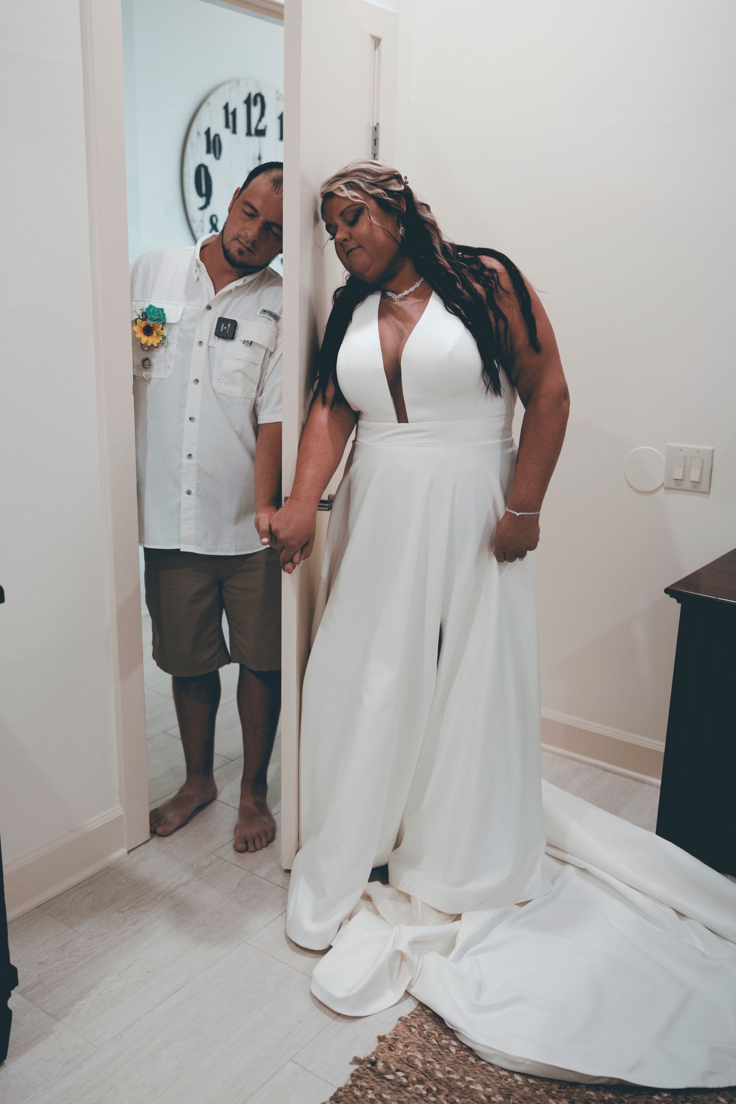 Two people pose together indoors, one wearing a white button-down shirt and shorts, the other in an elegant white jumpsuit.