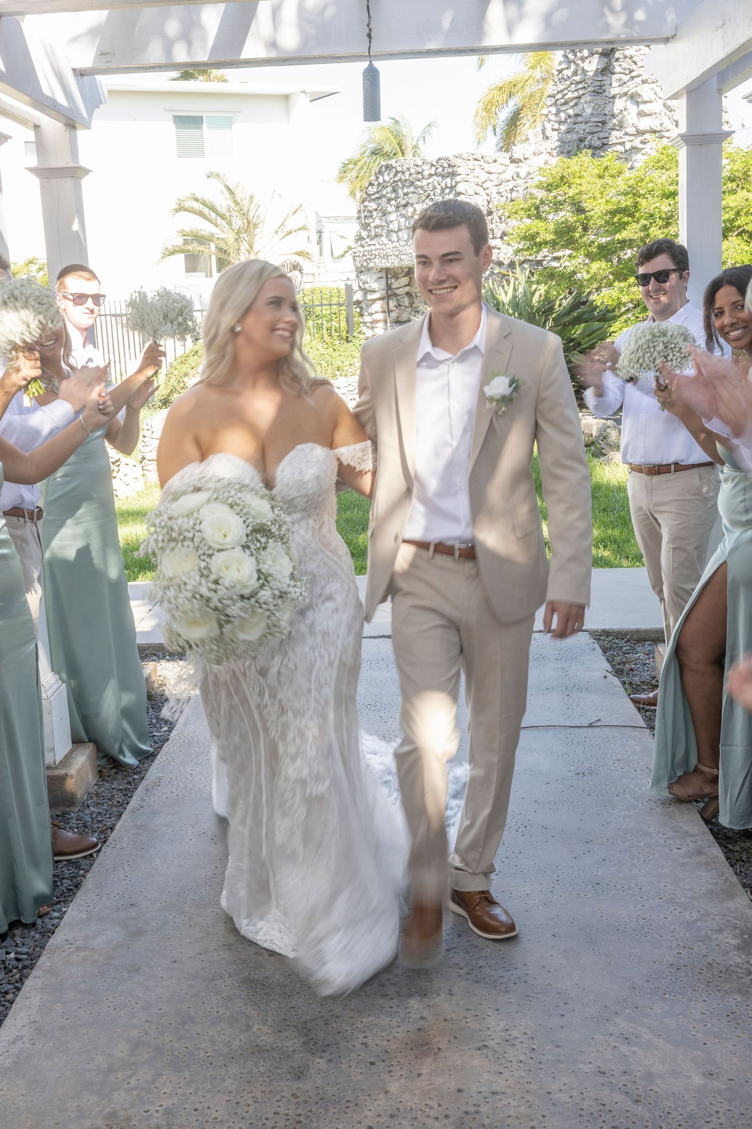 A couple walks down a covered pathway at their outdoor wedding while guests clap and celebrate in the background.