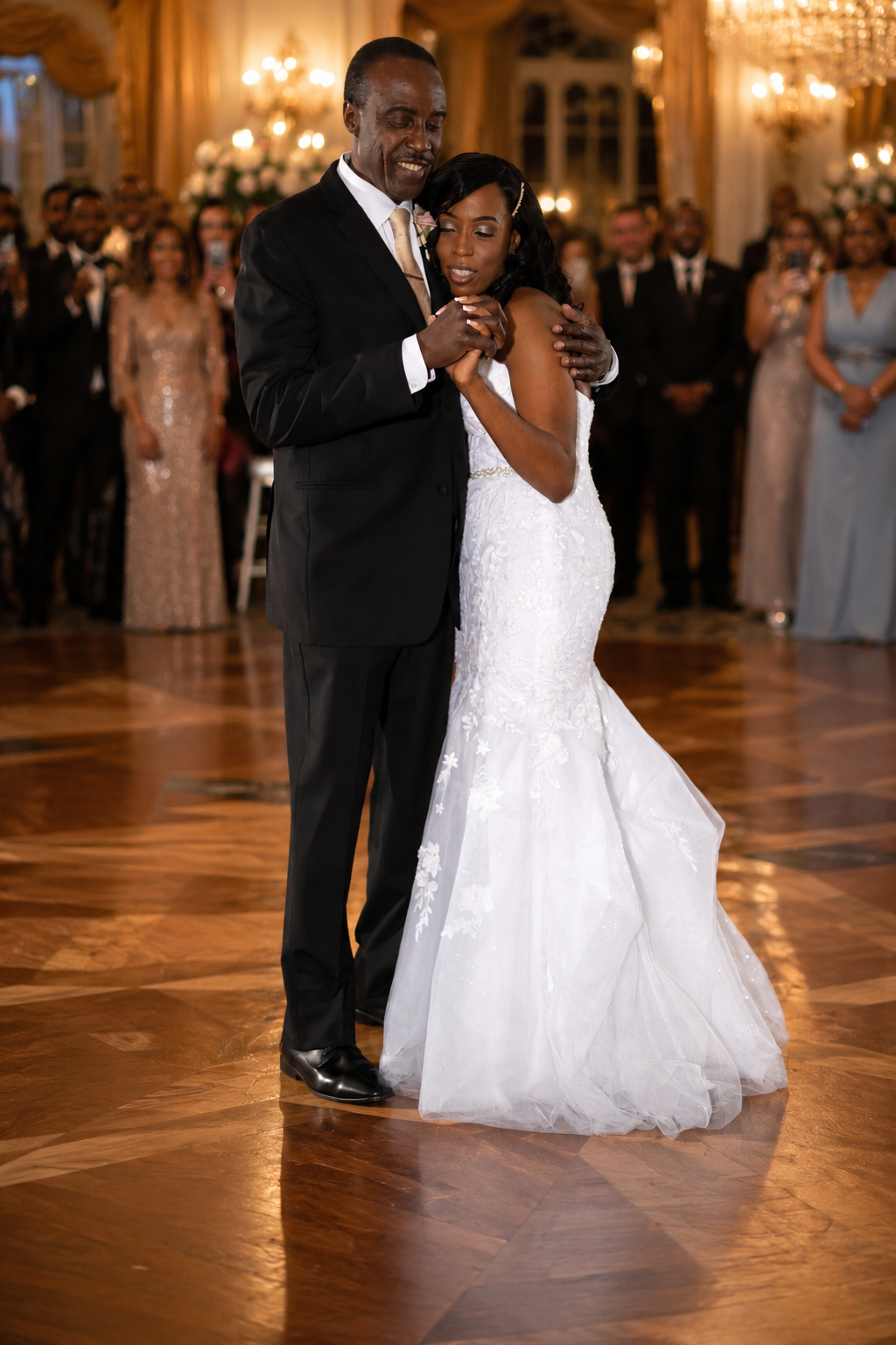 Bride and groom share their first dance in an elegant ballroom, guests watching as she wears a white mermaid gown.