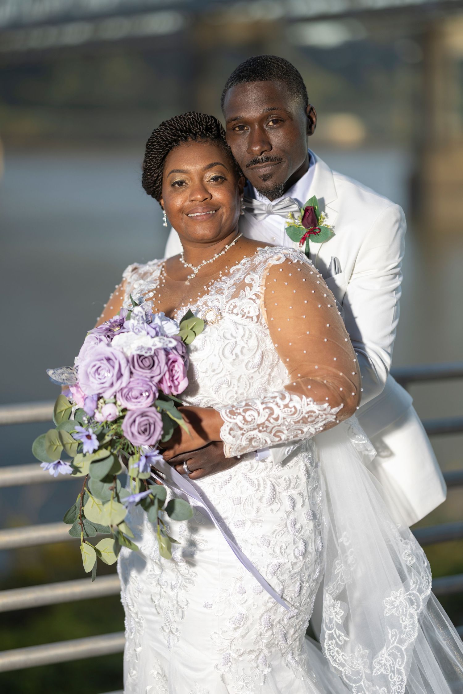 A Black bride and groom pose elegantly outdoors, she holds purple roses bouquet, he wears white tuxedo with floral boutonniere.