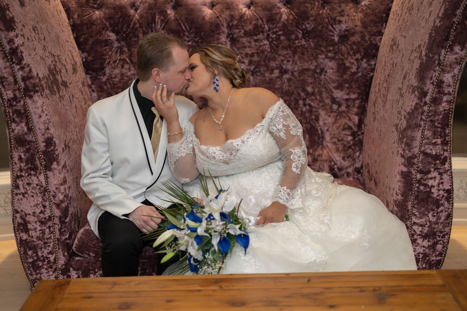 Bride and groom share a romantic kiss on a velvet chair, bride holding blue and white bouquet in wedding attire.