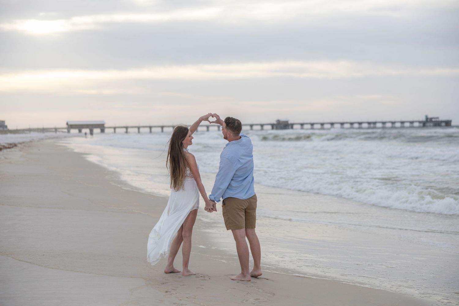 Couple holding hands and forming a heart shape on a sandy beach with a pier in the background at sunset.
