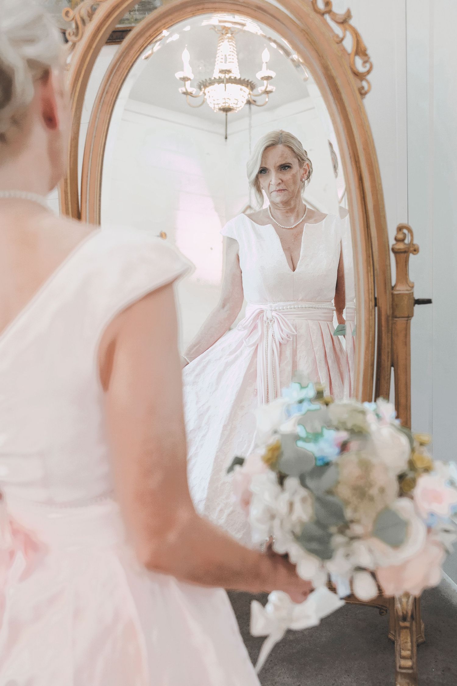 Bride in pink wedding dress holding floral bouquet gazes at her reflection in an ornate gold oval mirror.
