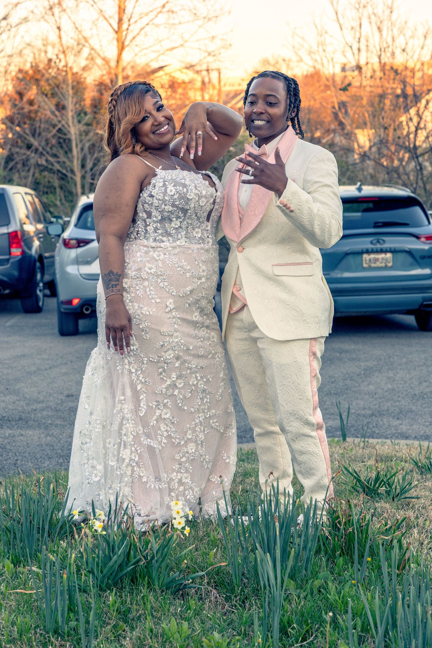 Couple in formal attire posing outdoors at sunset, woman in white gown and man in cream tuxedo.