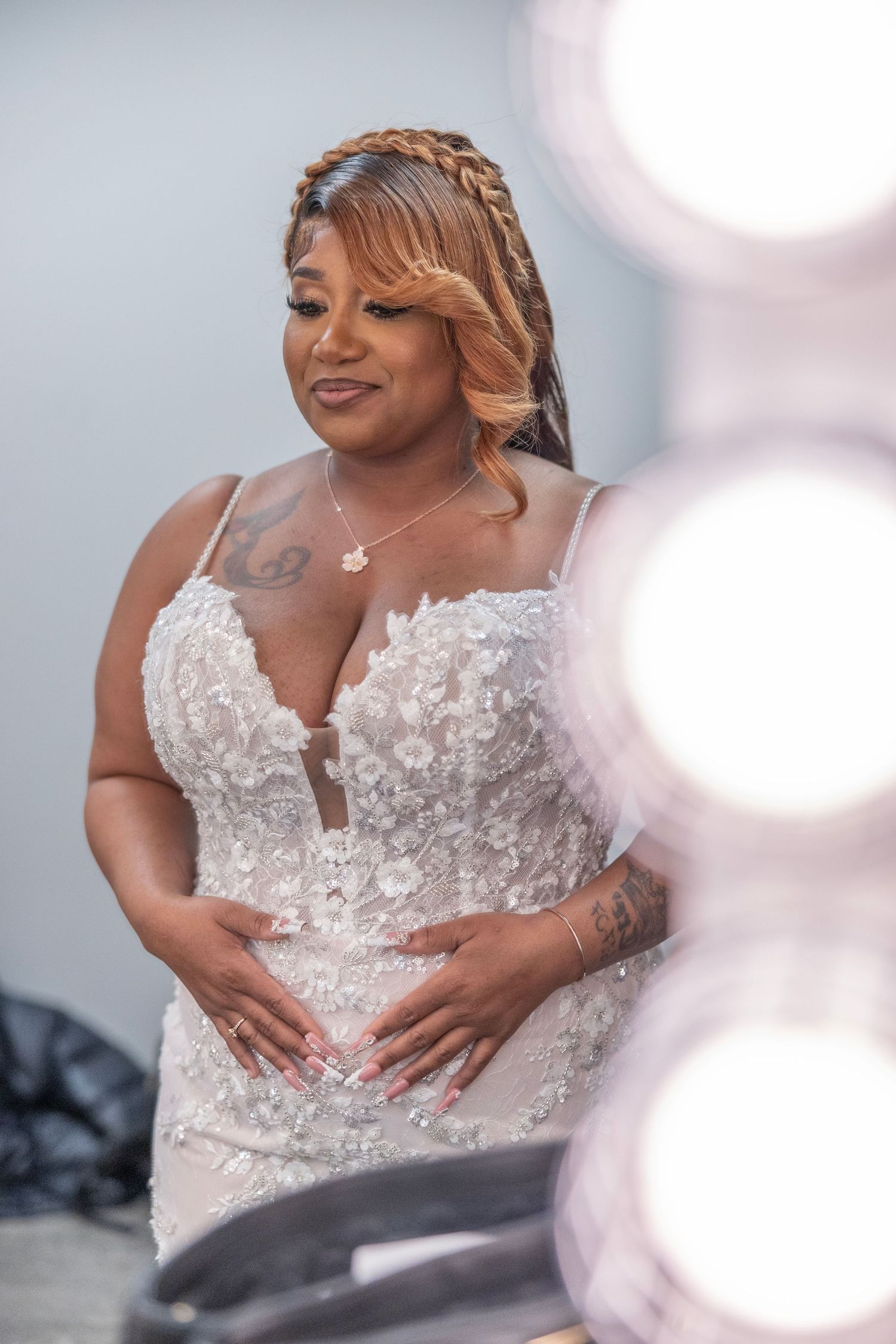 A bride in a lace wedding gown with floral appliques smiles softly, her braided updo and tattoos adding elegance.