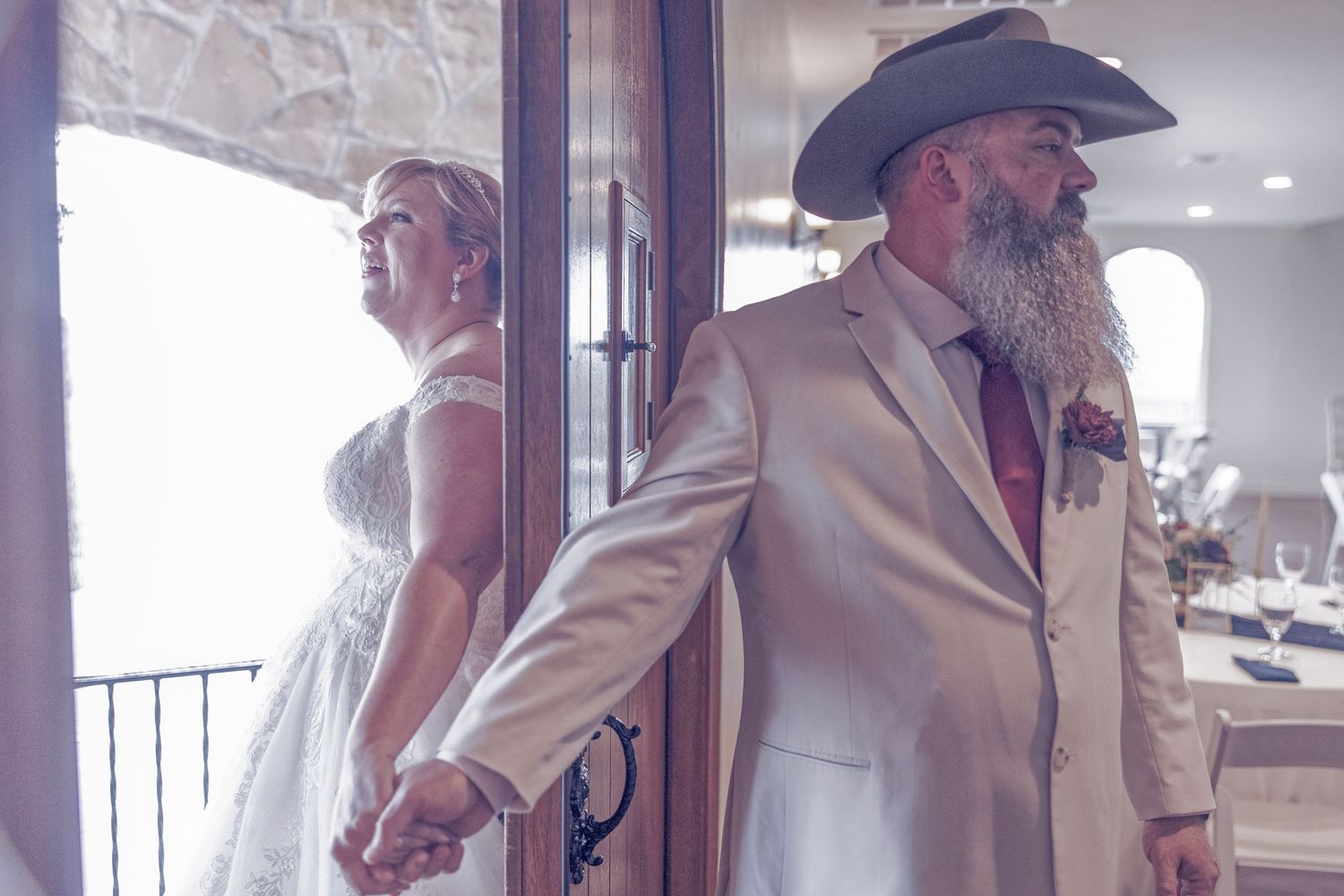 Bride and groom stand back-to-back holding hands before wedding ceremony, groom wearing white suit and cowboy hat with long beard.