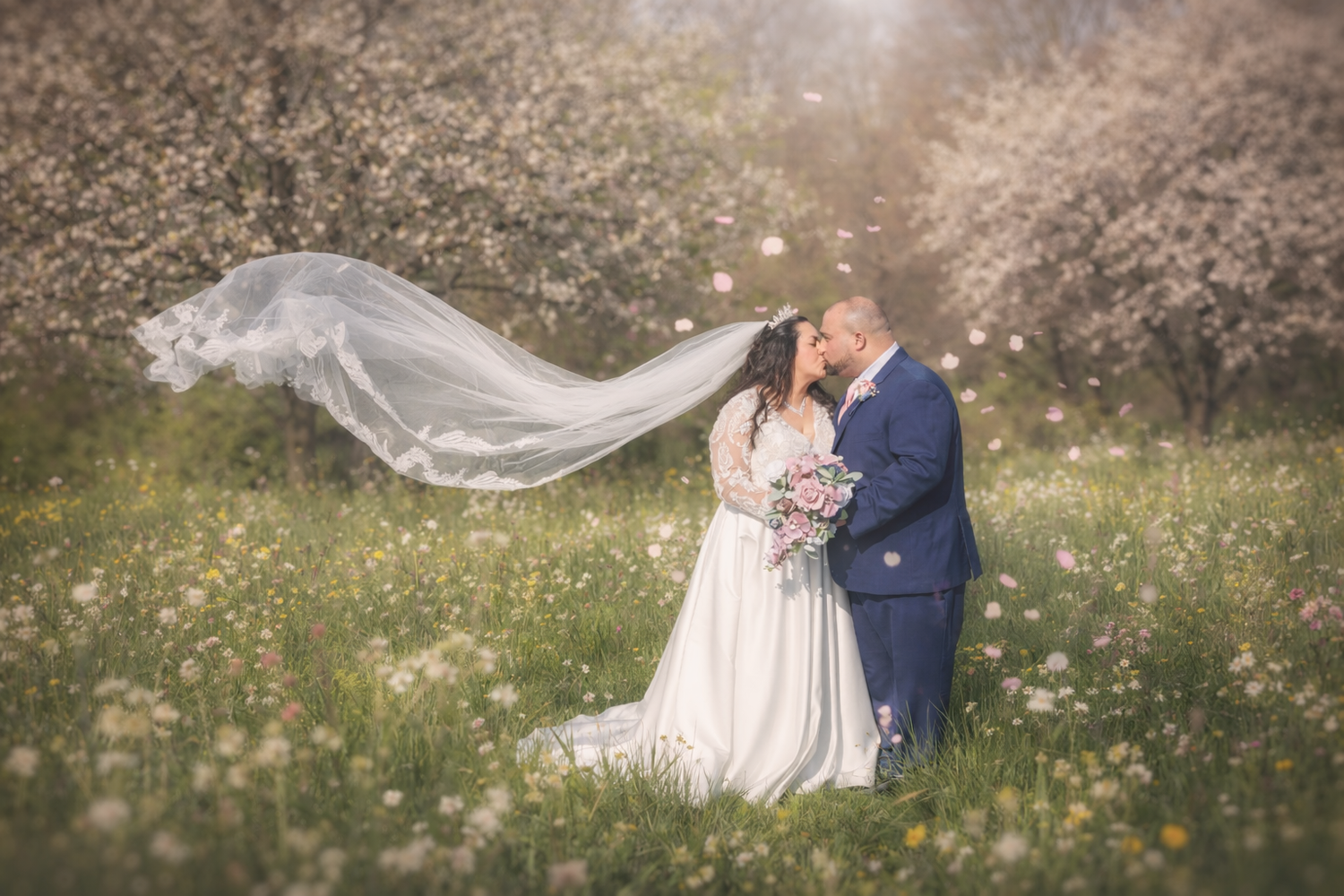 Bride and groom kissing in a blooming meadow, wedding veil flowing in the wind, surrounded by spring cherry blossom trees.