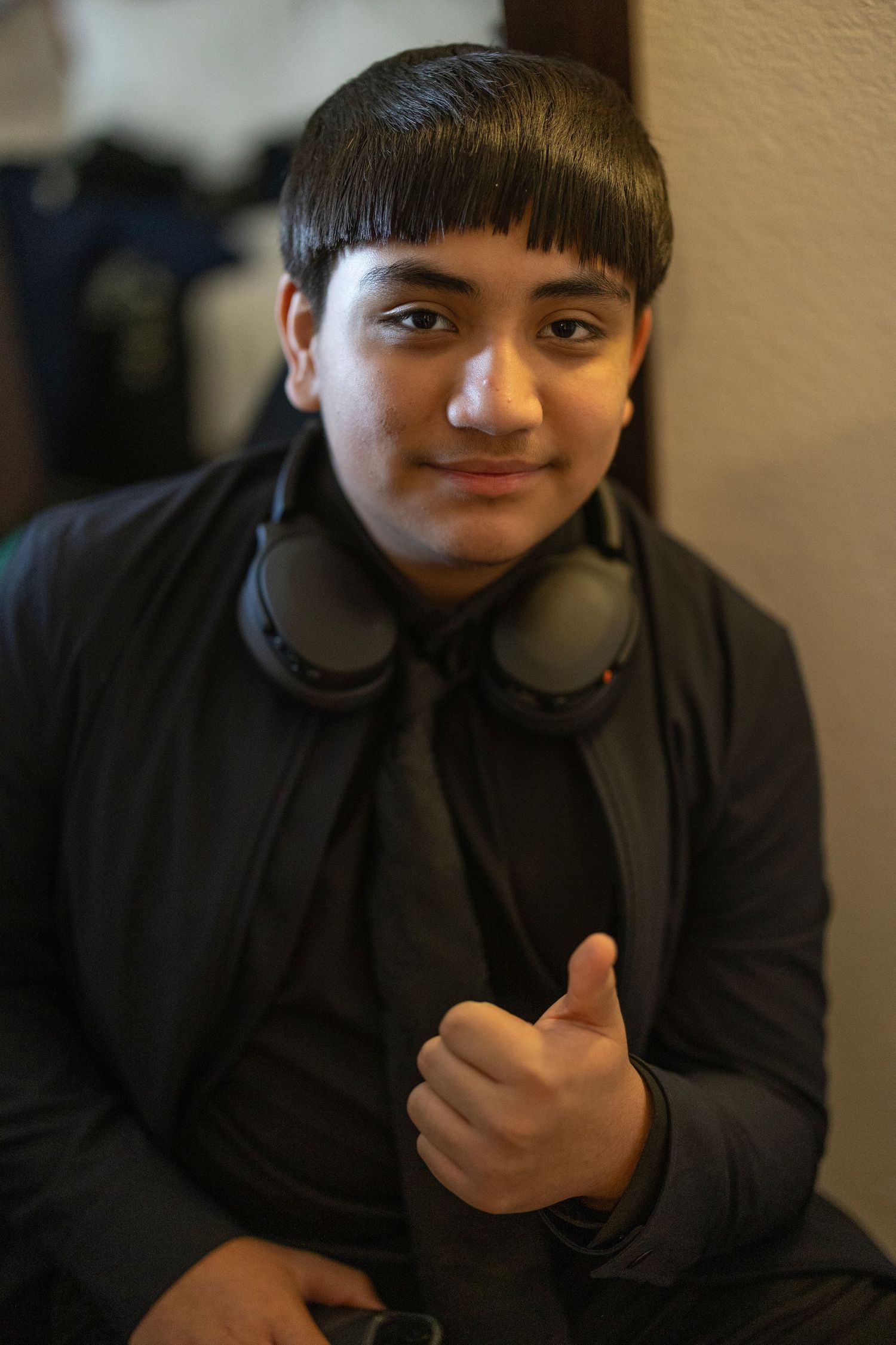 Young teen with bowl cut wearing black jacket and headphones around neck giving thumbs up gesture indoors.