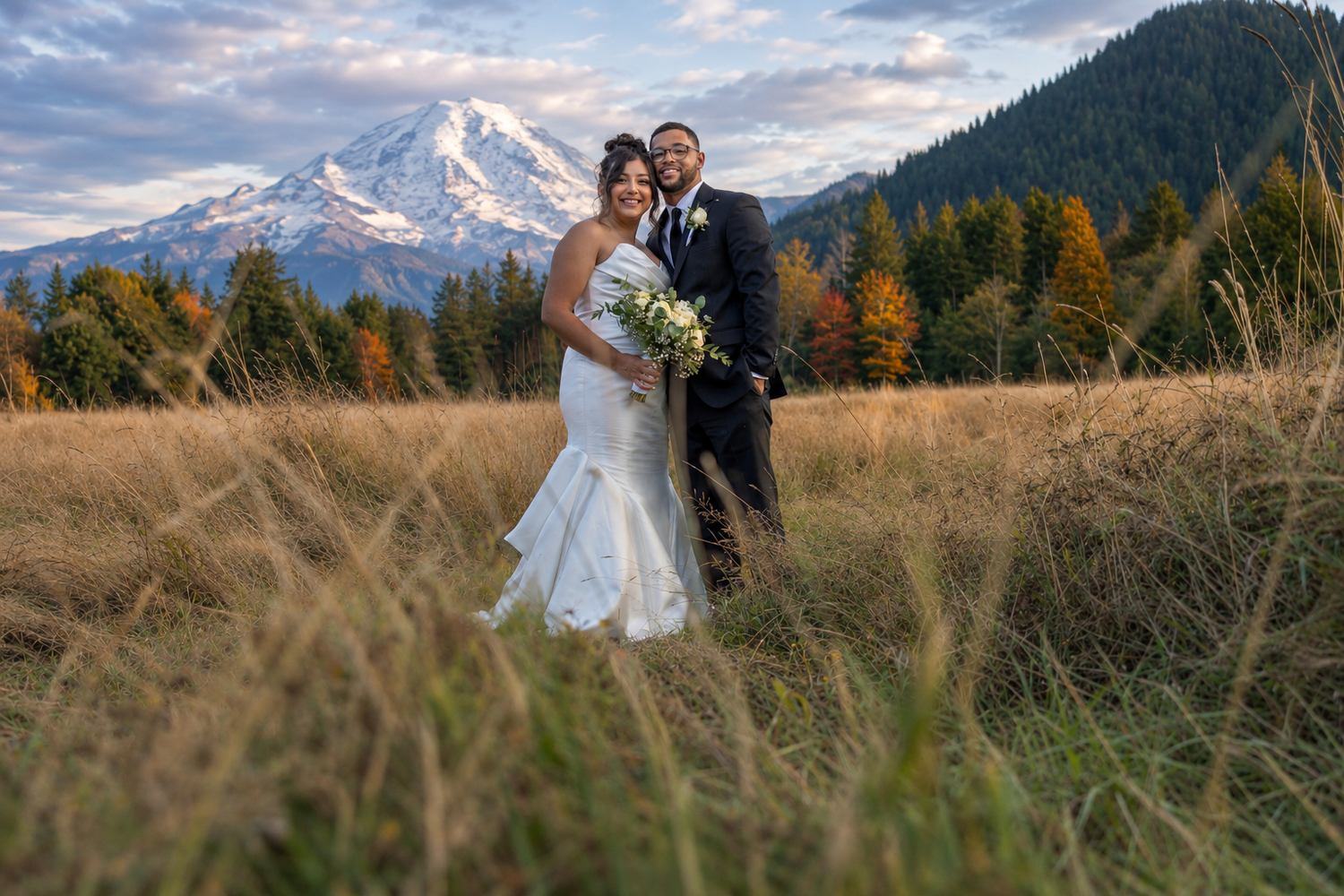 Bride and groom pose in golden meadow with snow-capped mountain and autumn trees backdrop.