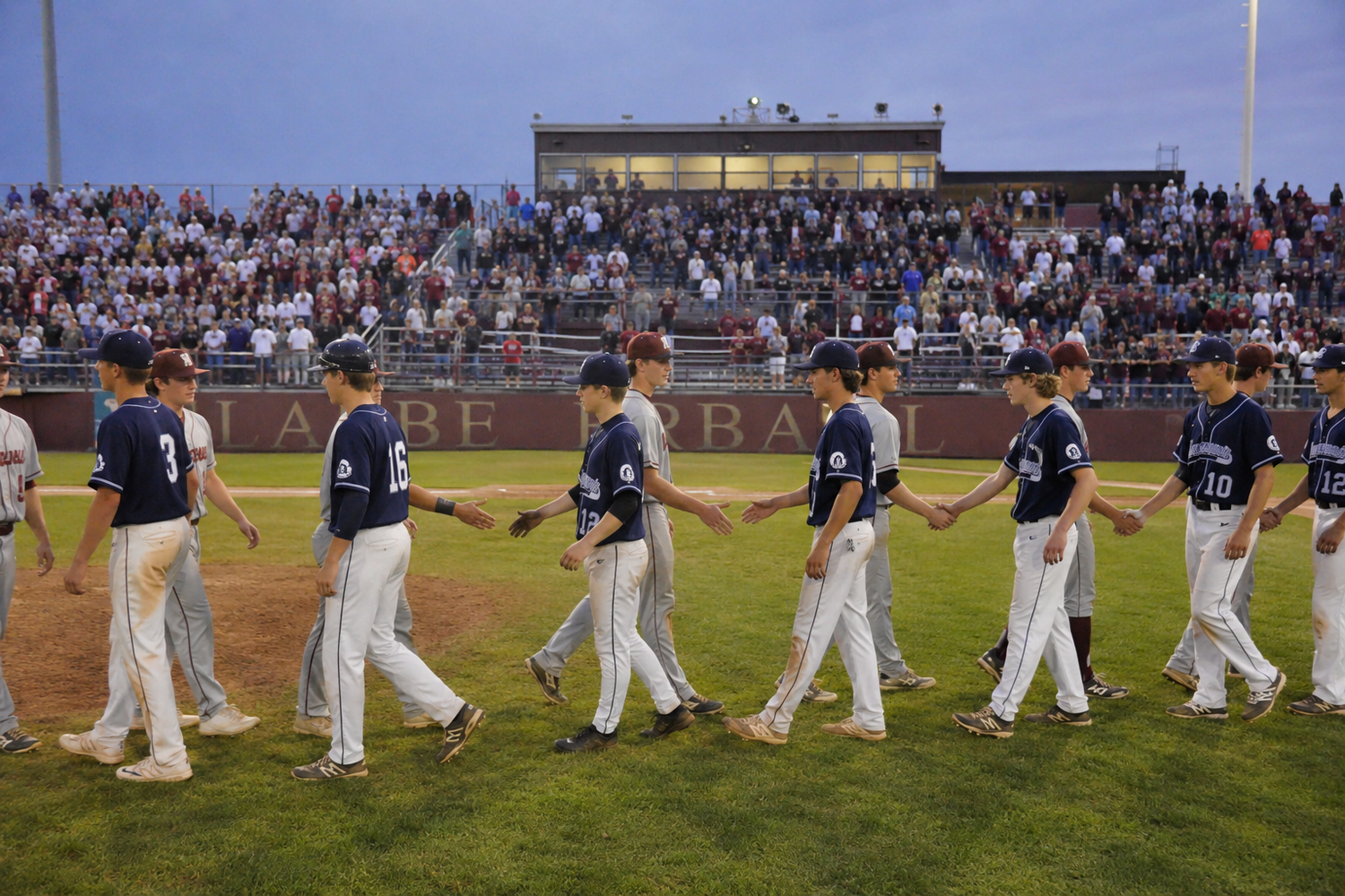 Baseball players shake hands on the field after a game in front of a packed stadium crowd at dusk.
