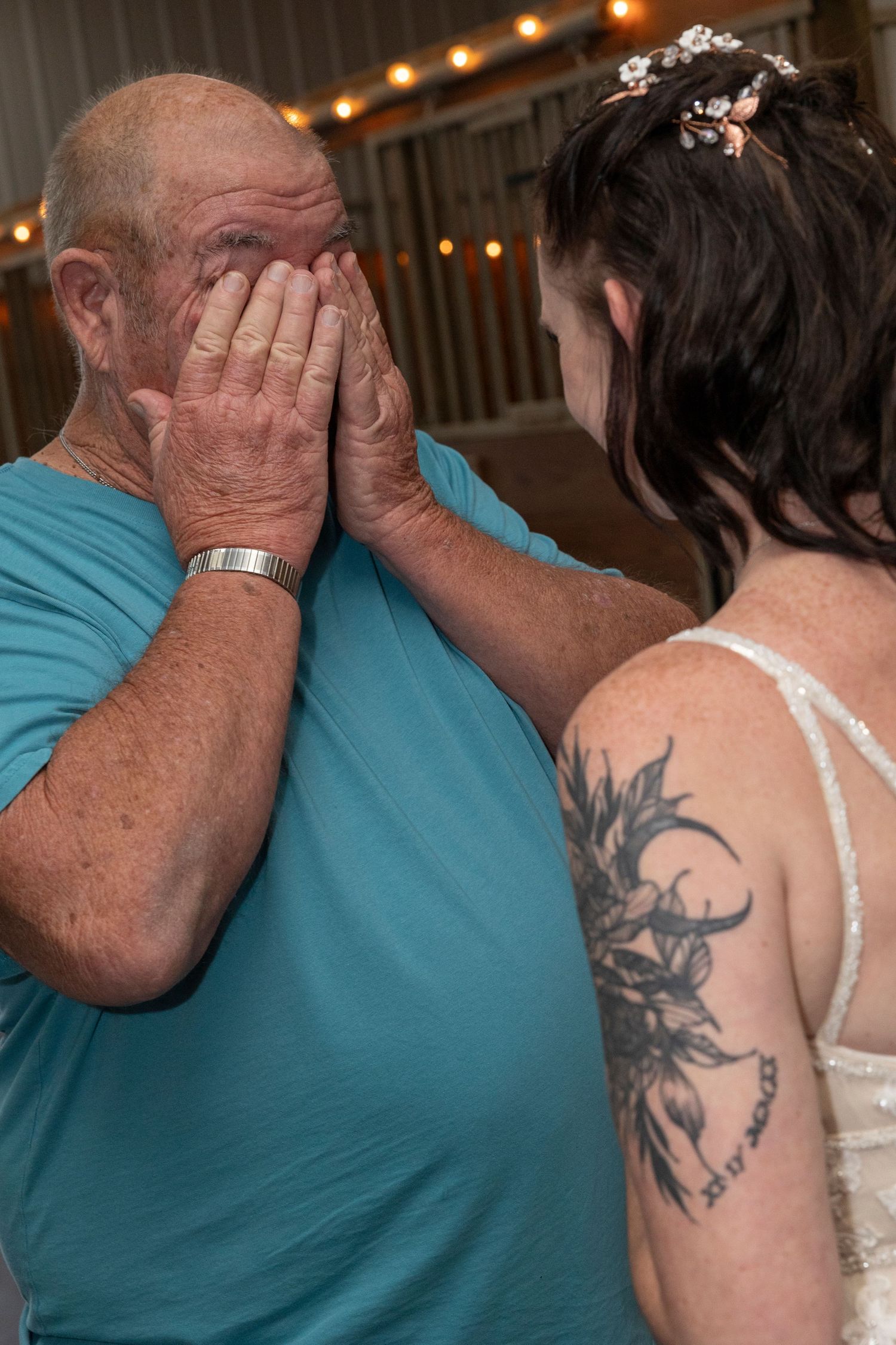 An emotional moment between someone in a teal shirt and a person in white with a visible floral tattoo at a wedding.