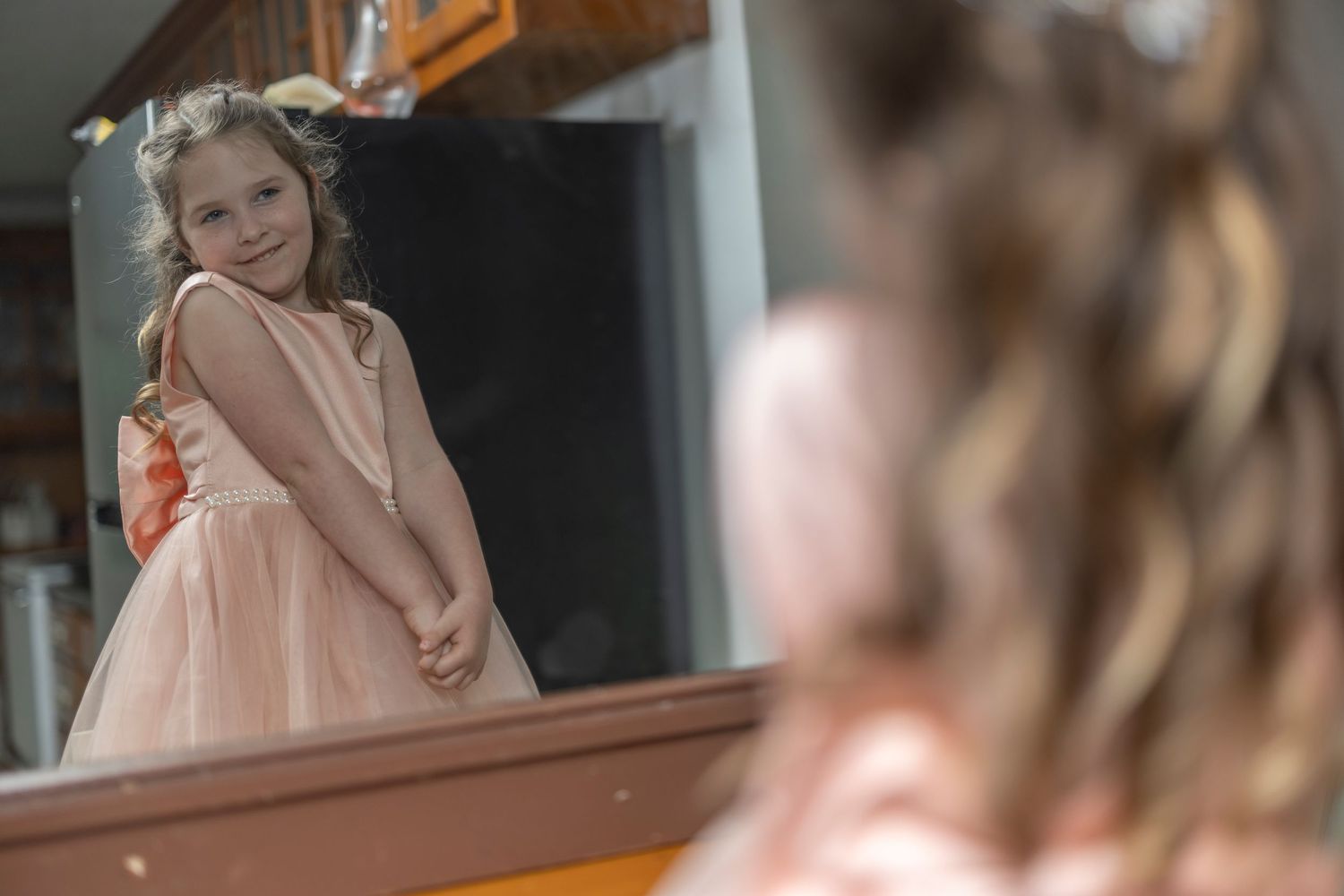 A youth in a peach-colored dress smiles while looking at their reflection in a mirror.