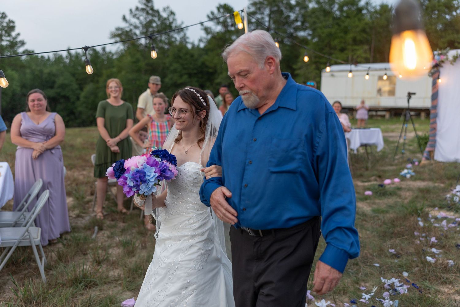 Bride walks down outdoor aisle with older man in blue shirt, holding purple bouquet, guests watching at rustic wedding.