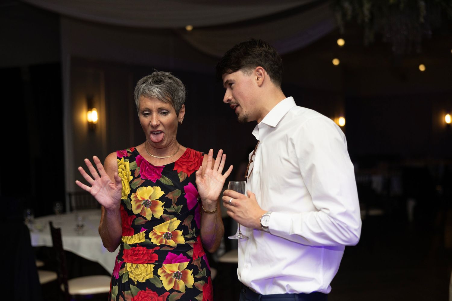 A woman in a floral dress and a young man in a white shirt converse at an elegant indoor event.