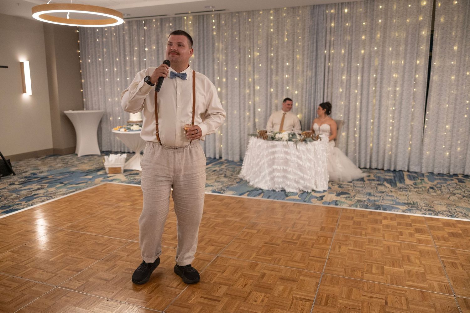 Man giving a speech at a wedding reception with bride and groom seated at the head table in the background.