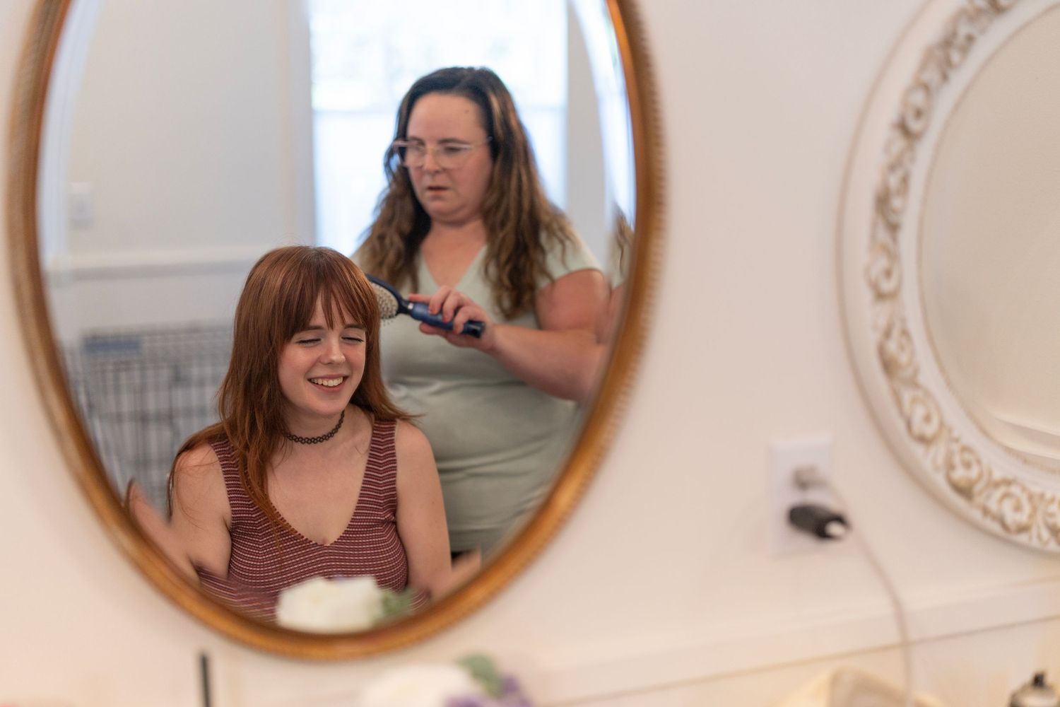 Woman styling red-haired girl's hair with brush, smiling reflection visible in oval bathroom mirror.