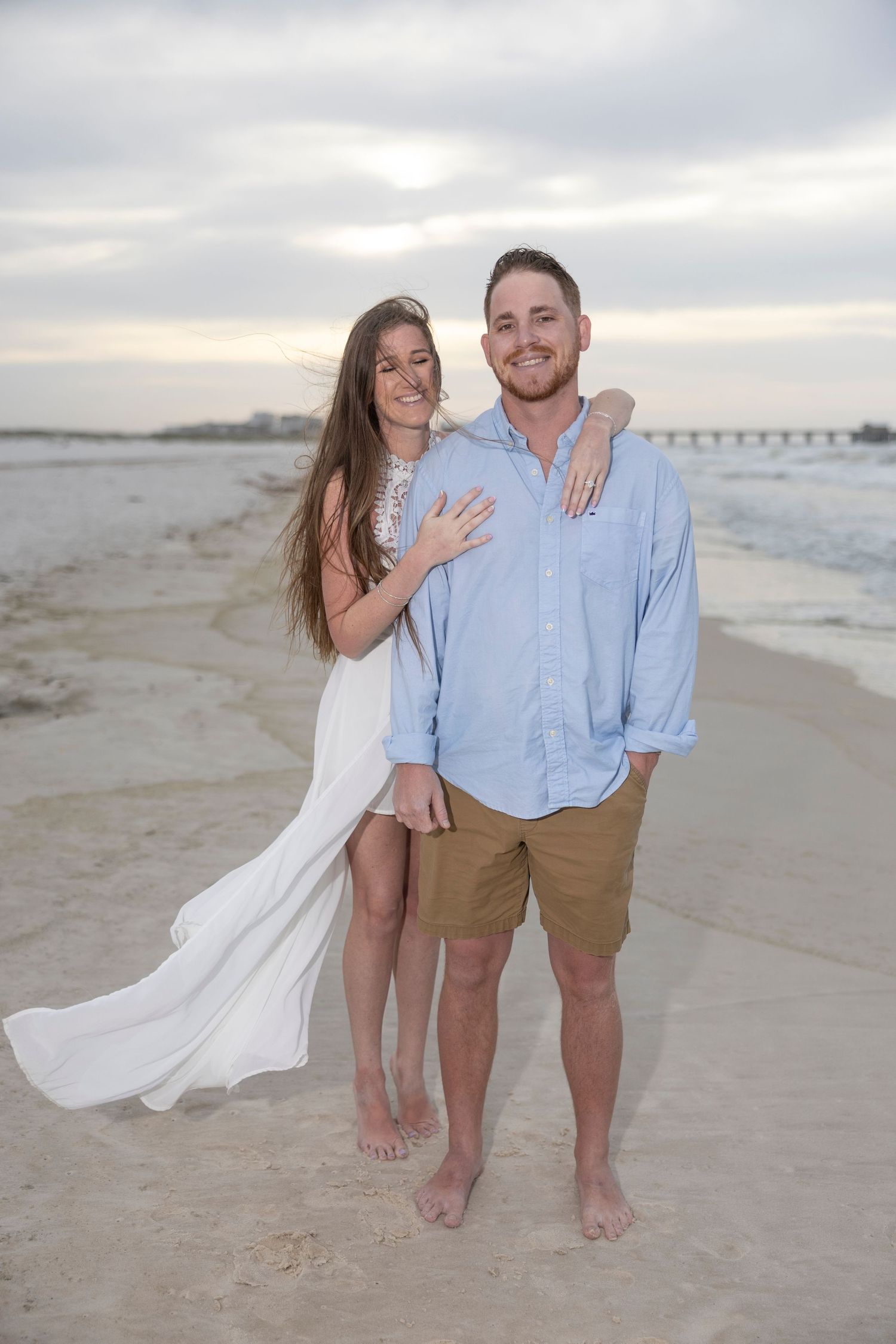 Couple posing together on a sandy beach at sunset, woman in white dress and man in blue shirt and khaki shorts.