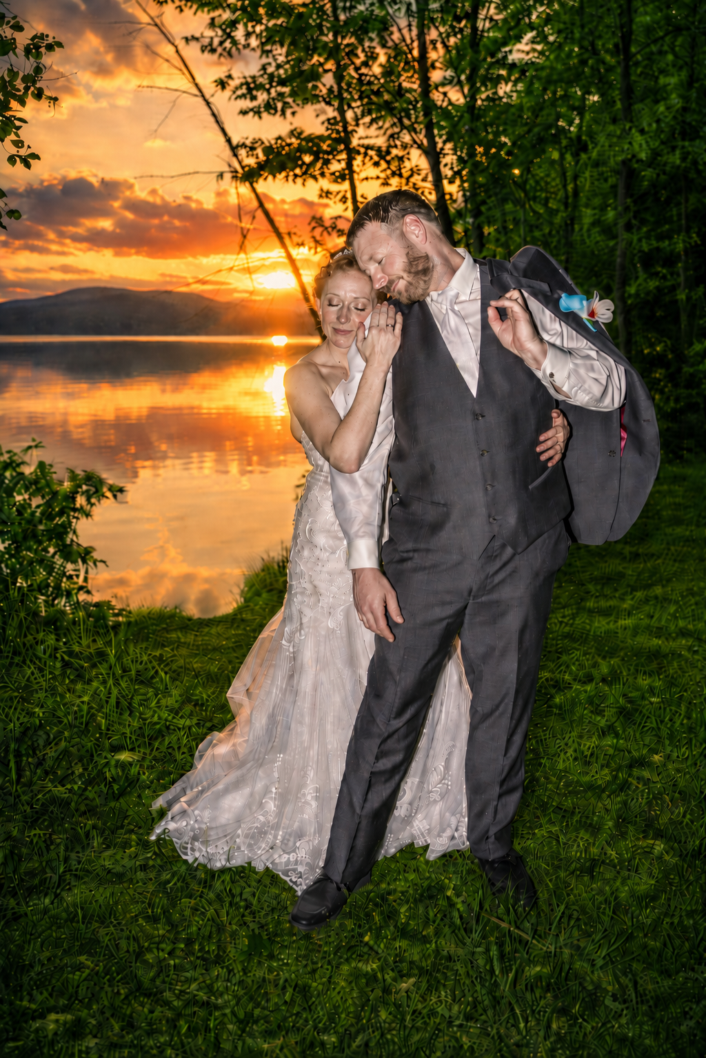 Newlywed couple embracing at sunset by a serene lake, bride in lace gown and groom in gray suit, golden sky reflected on water.