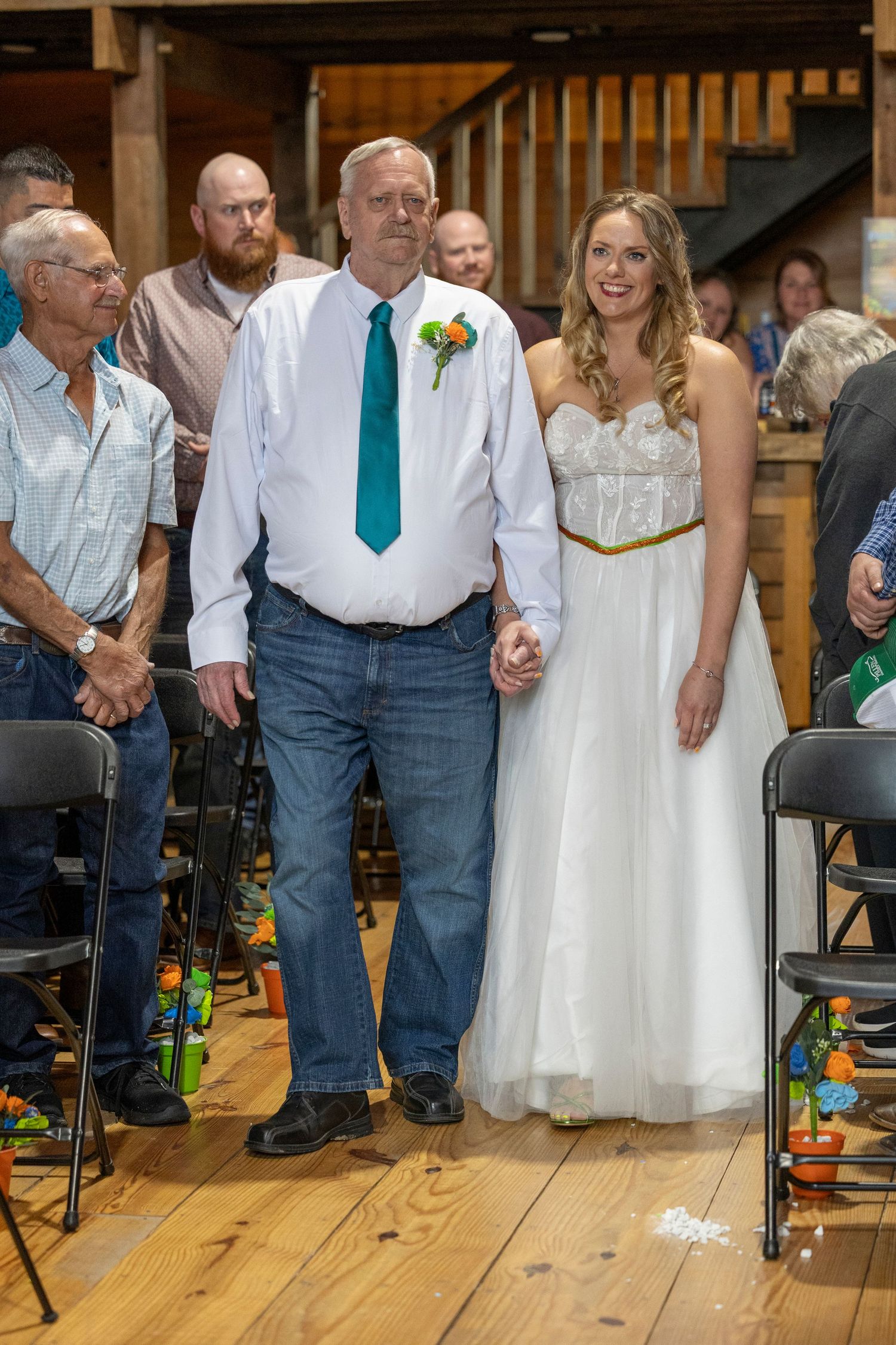 Bride in white gown walks down aisle with father in teal tie at rustic barn wedding ceremony.