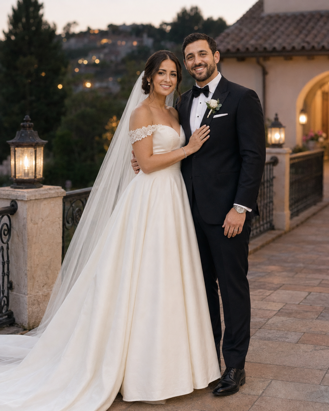 A bride in an off-shoulder white ballgown with veil and groom in black tuxedo pose together at an outdoor wedding venue.