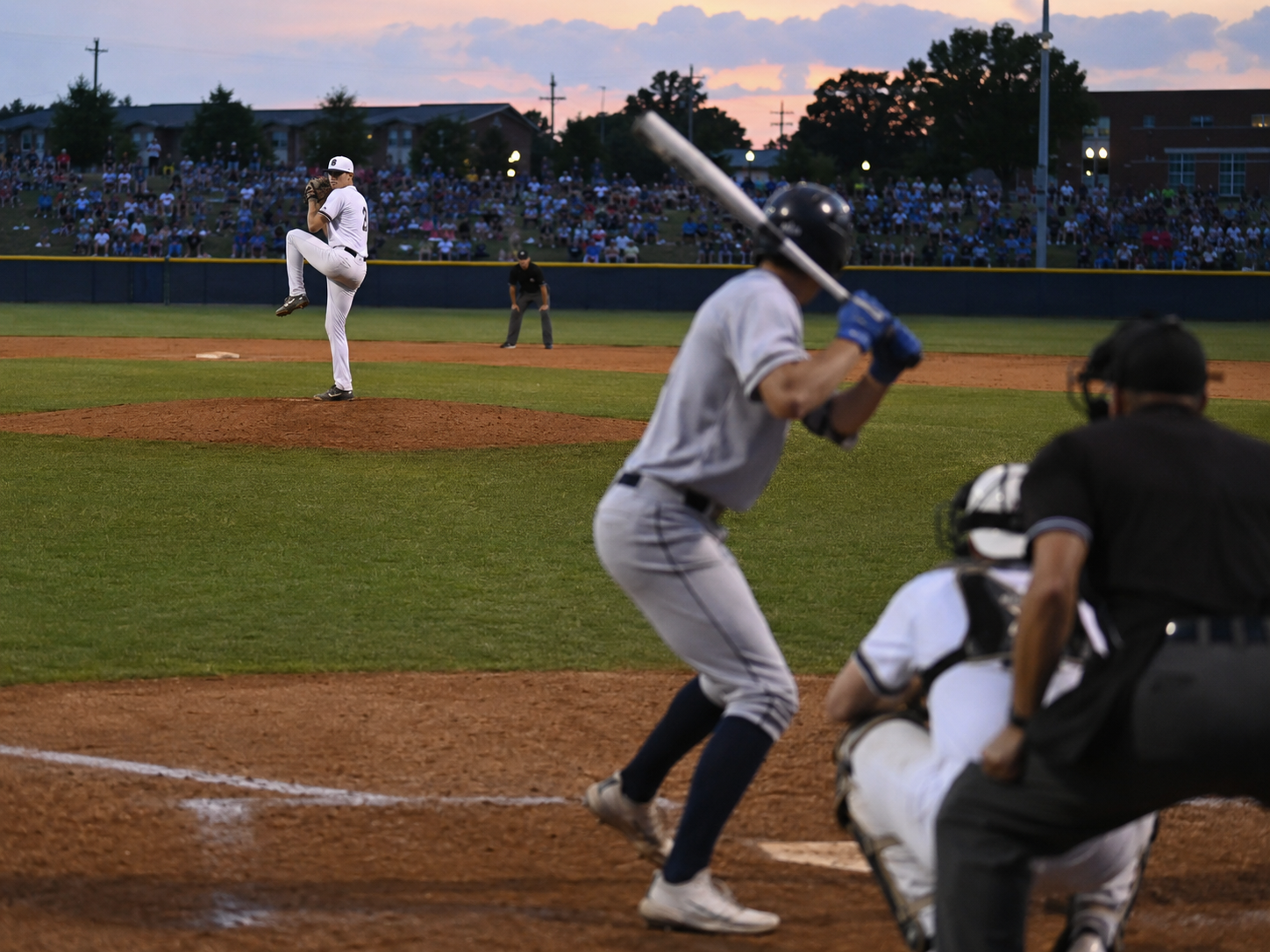 Baseball batter prepares to swing as pitcher winds up at dusk in a packed minor league stadium.