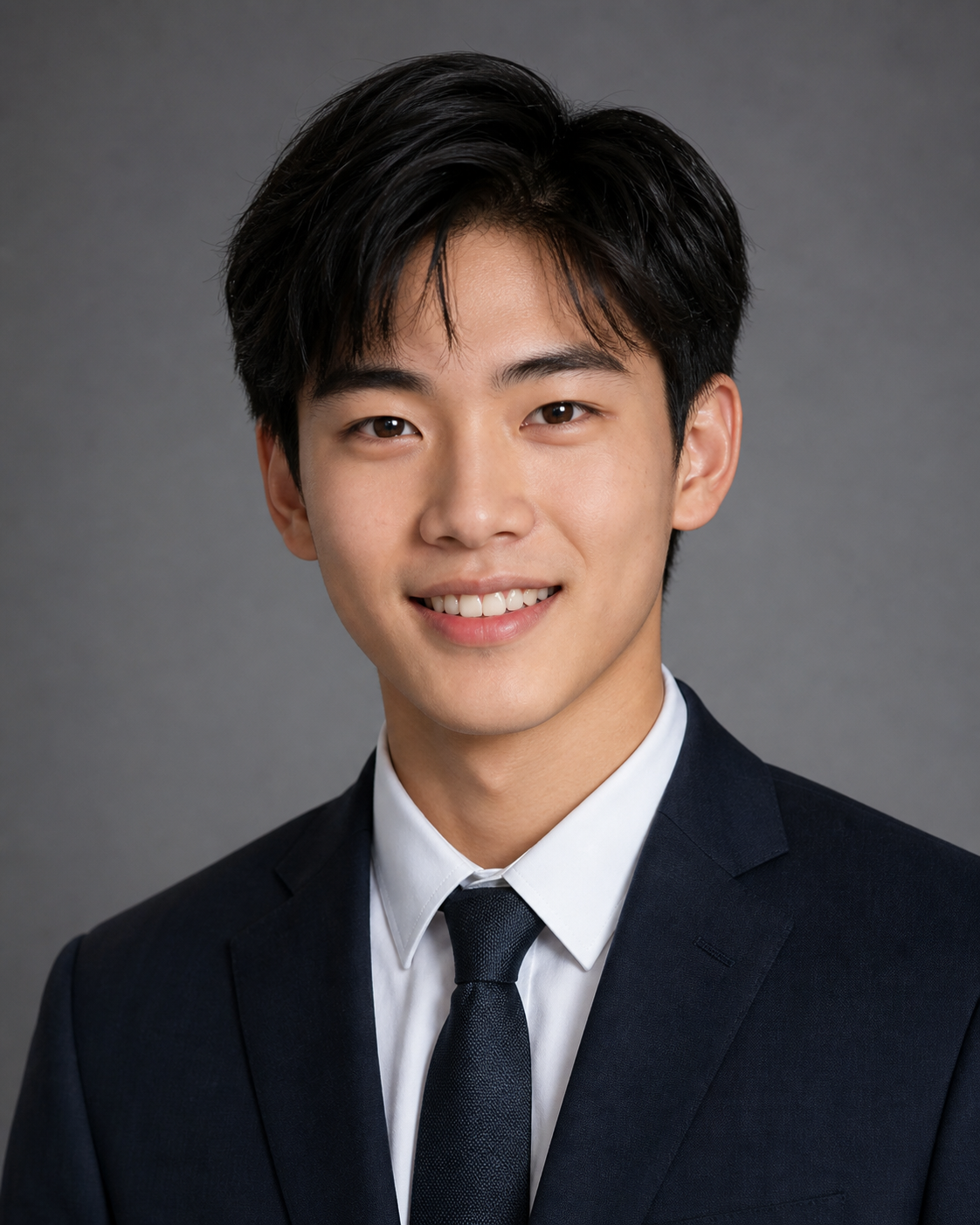 Young man in navy suit and tie smiling professionally against gray background in formal headshot photo.