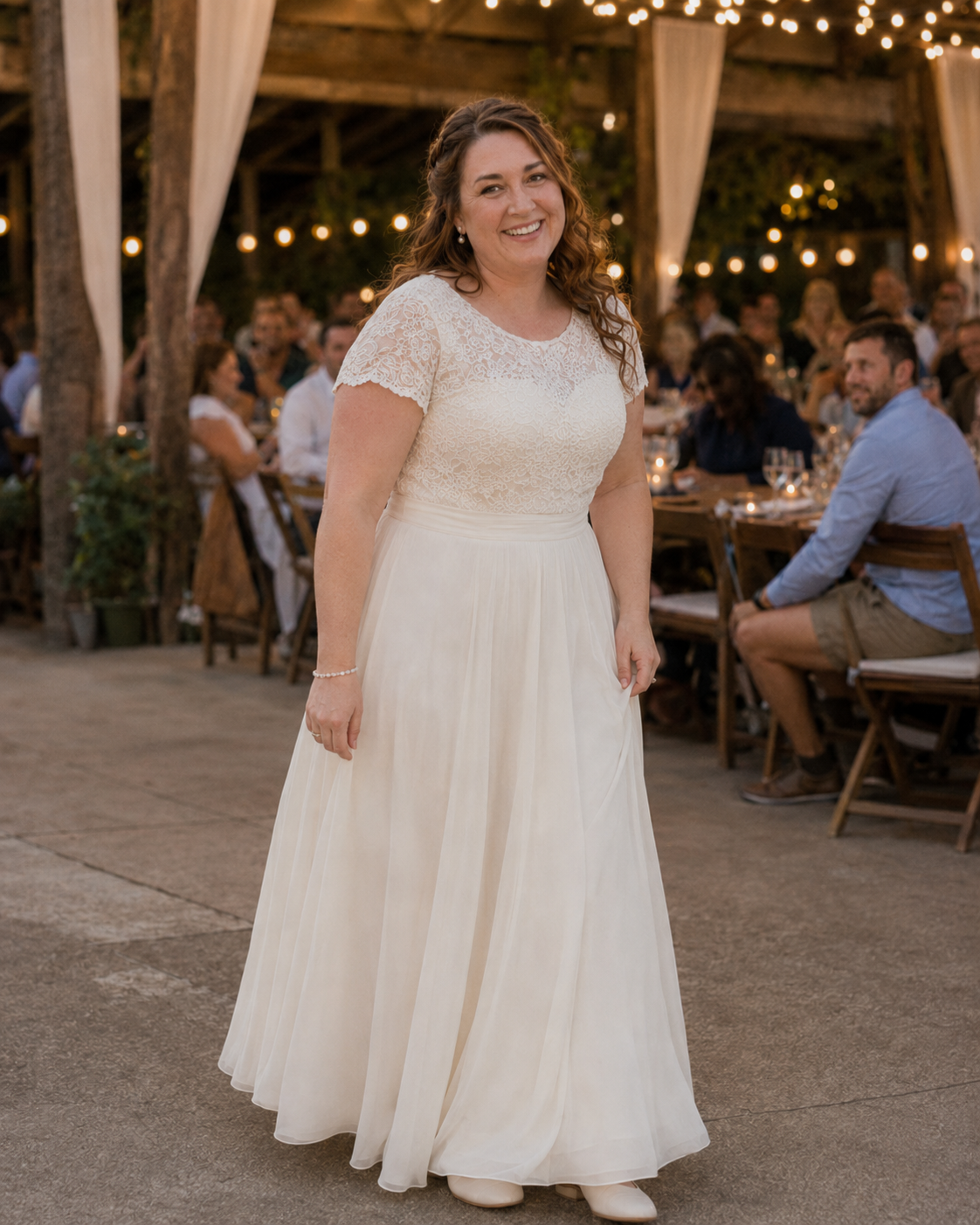 Bride in white lace short-sleeve wedding gown smiling at an outdoor rustic wedding reception.