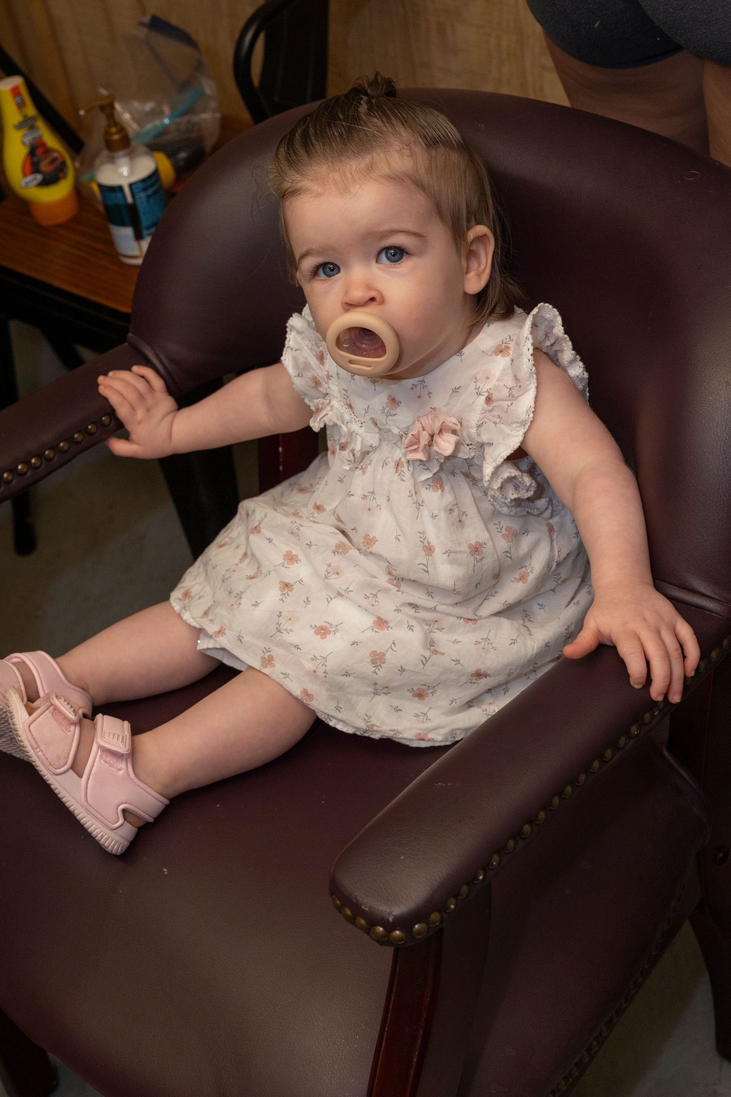 Baby in a floral dress and pink shoes sits in a large brown leather armchair with surprised expression.