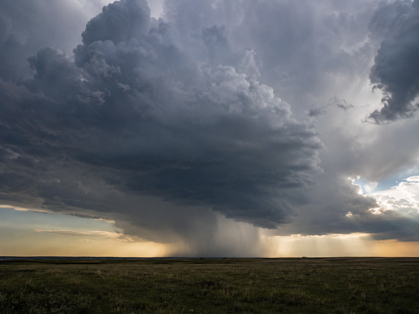 Dramatic storm cell over vast green plains at sunset with heavy rain curtain falling beneath dark clouds.
