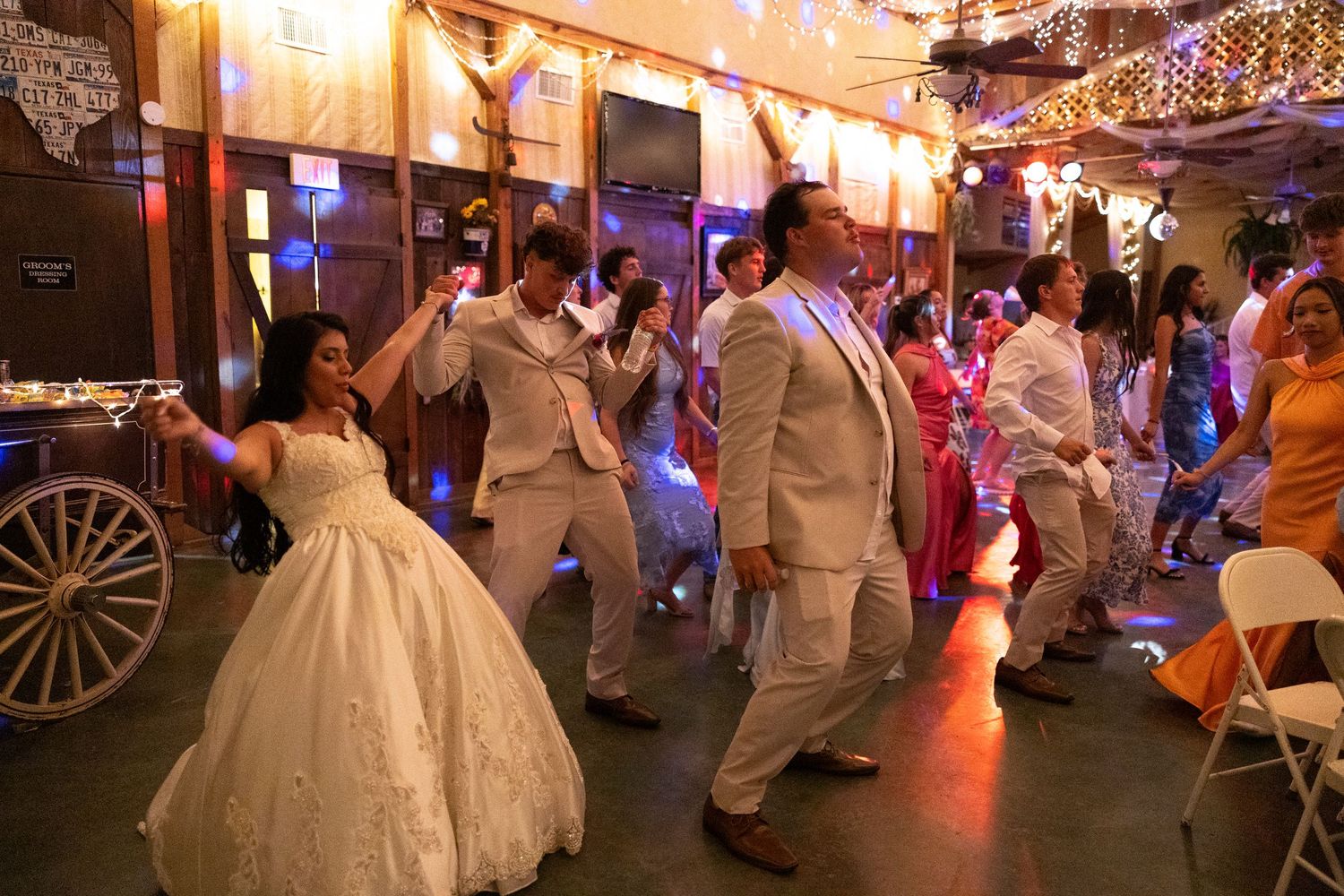 Bride and groom dancing with guests at a lively wedding reception in a decorated rustic venue.